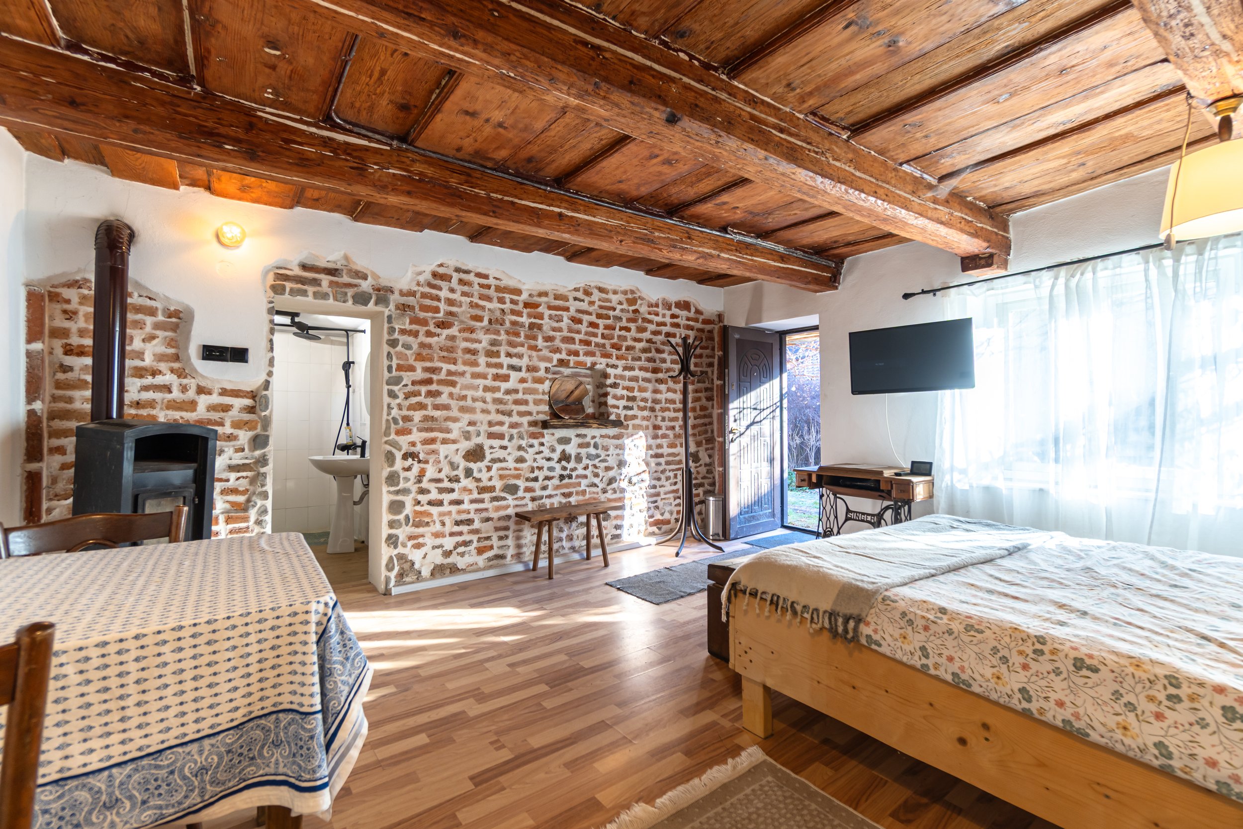 A cozy bedroom with a rustic wooden ceiling, exposed brick wall, wooden flooring, a bed with floral sheets, a wall-mounted TV, a small table or desk by the window, a coat rack, a bench near the brick wall, and a doorway leading to a bathroom with a s
