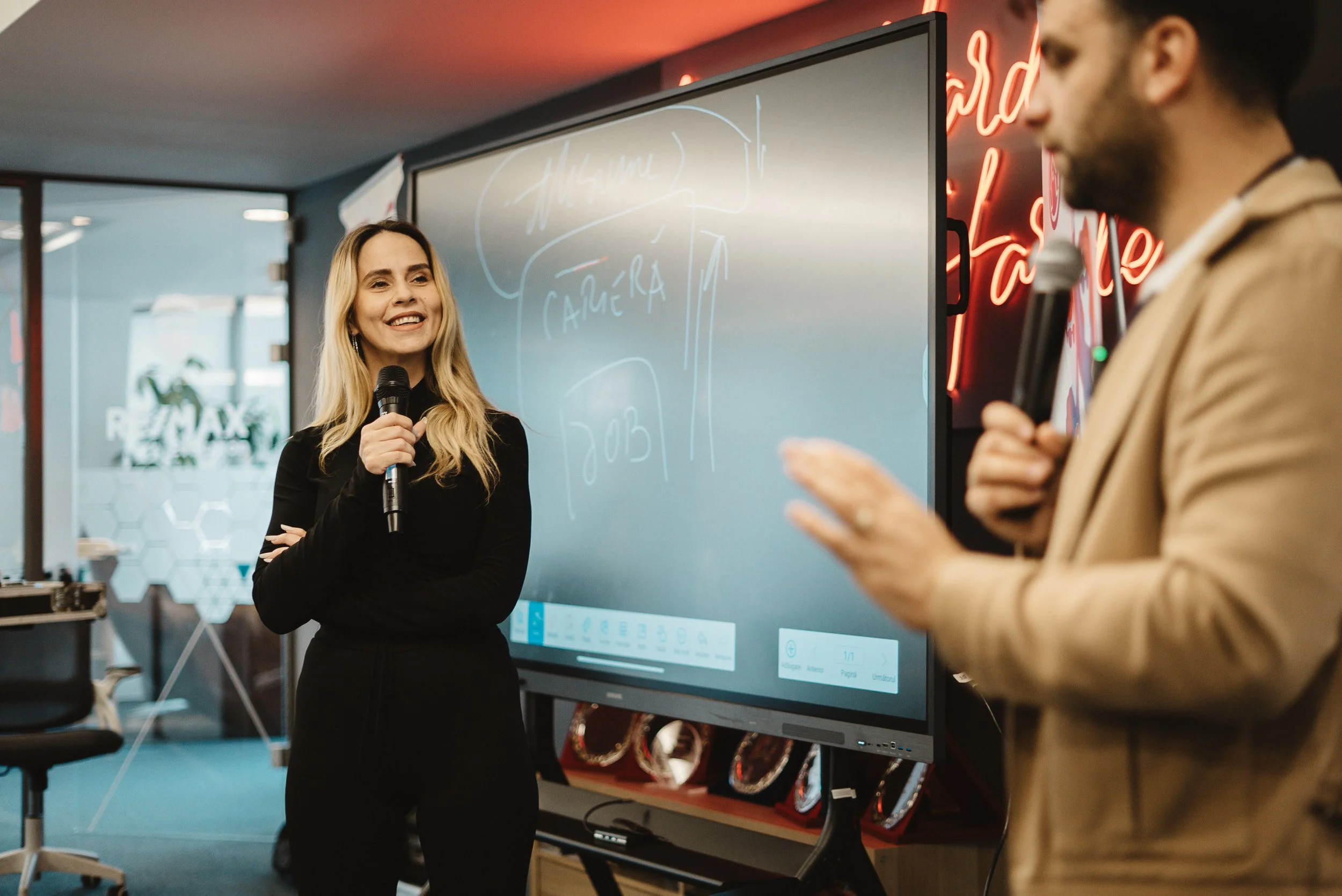 A woman with long blonde hair, wearing a black outfit, smiling and holding a microphone, standing in front of a large digital screen during a presentation. In the background are modern office elements and a neon sign.