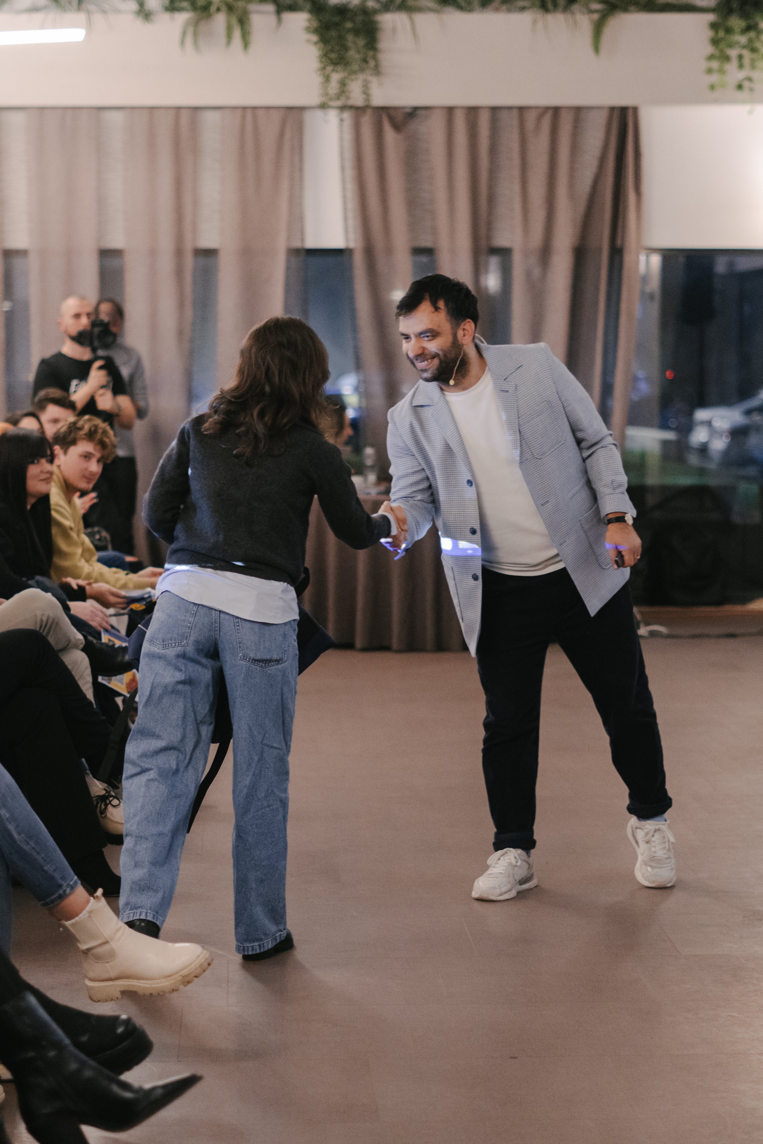 A man in a light gray blazer and white sneakers is shaking hands with a woman in casual jeans and a black jacket in a room with an audience. The woman has her back to the camera, and other people are seated on the left. Two people in the background a