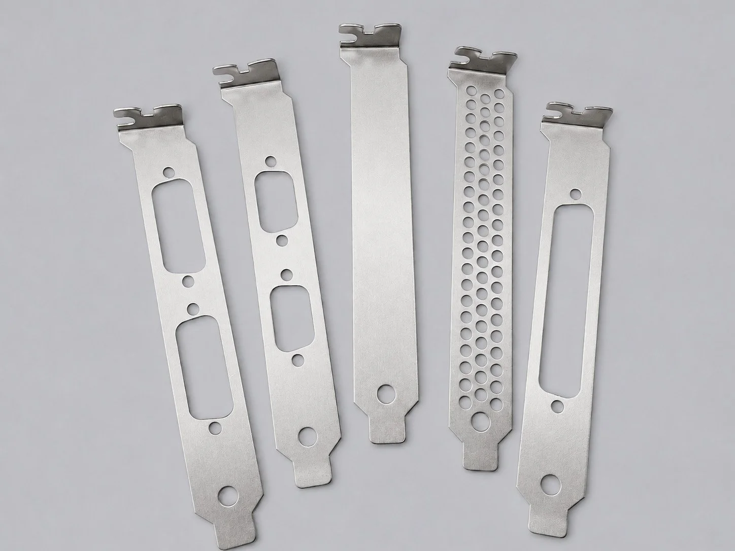 Five metal surgical blades laid out flat on a light gray surface, each with different hole patterns and cutouts for use in medical or cosmetic procedures.