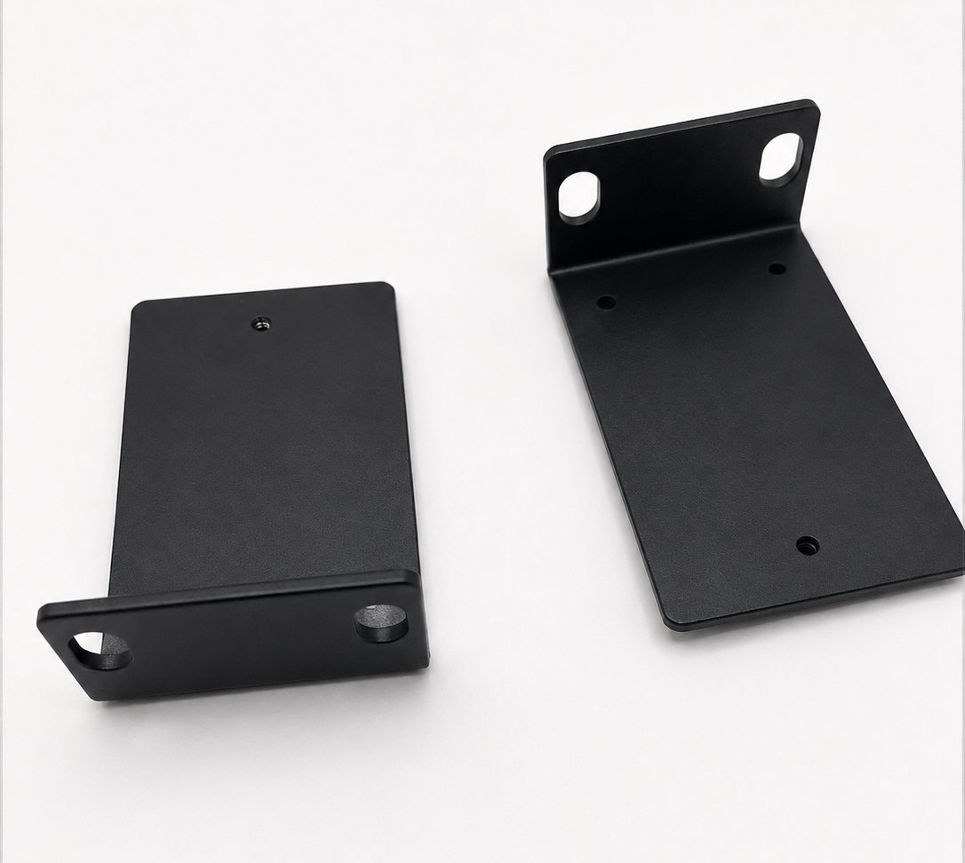 Two black metal mounting brackets on a white surface, one with side walls and the other with a vertical support and holes for screws.