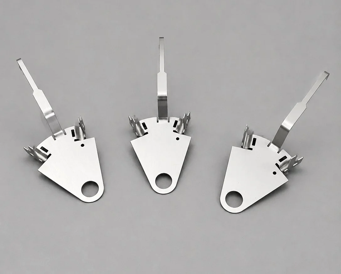 Three metallic mechanical parts with flat bases and protruding prongs, arranged on a gray background.