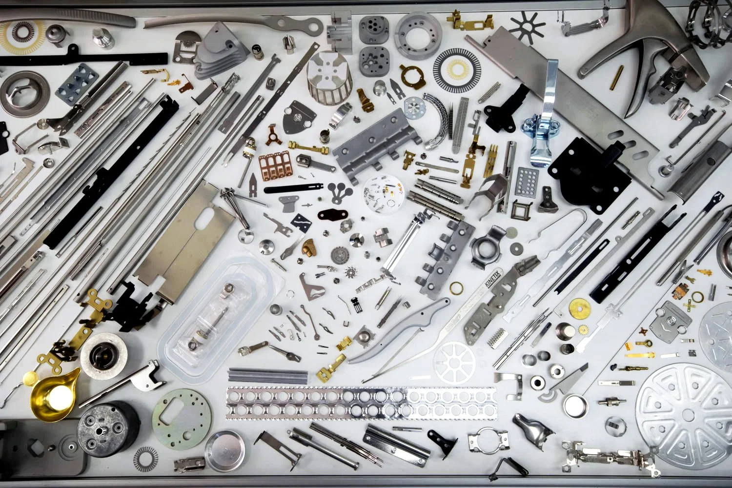 A collection of watch parts and gears spread across a white surface, including metal rods, gears, springs, screws, and other small components used in watchmaking.