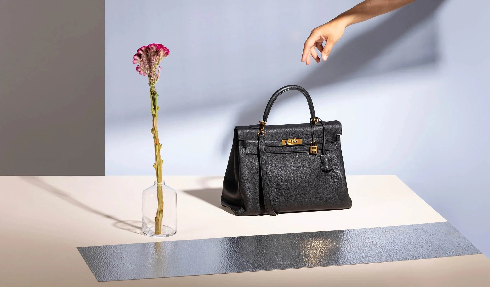 A black handbag on a table with a glass vase containing a pink flower, and a hand reaching towards the handbag.