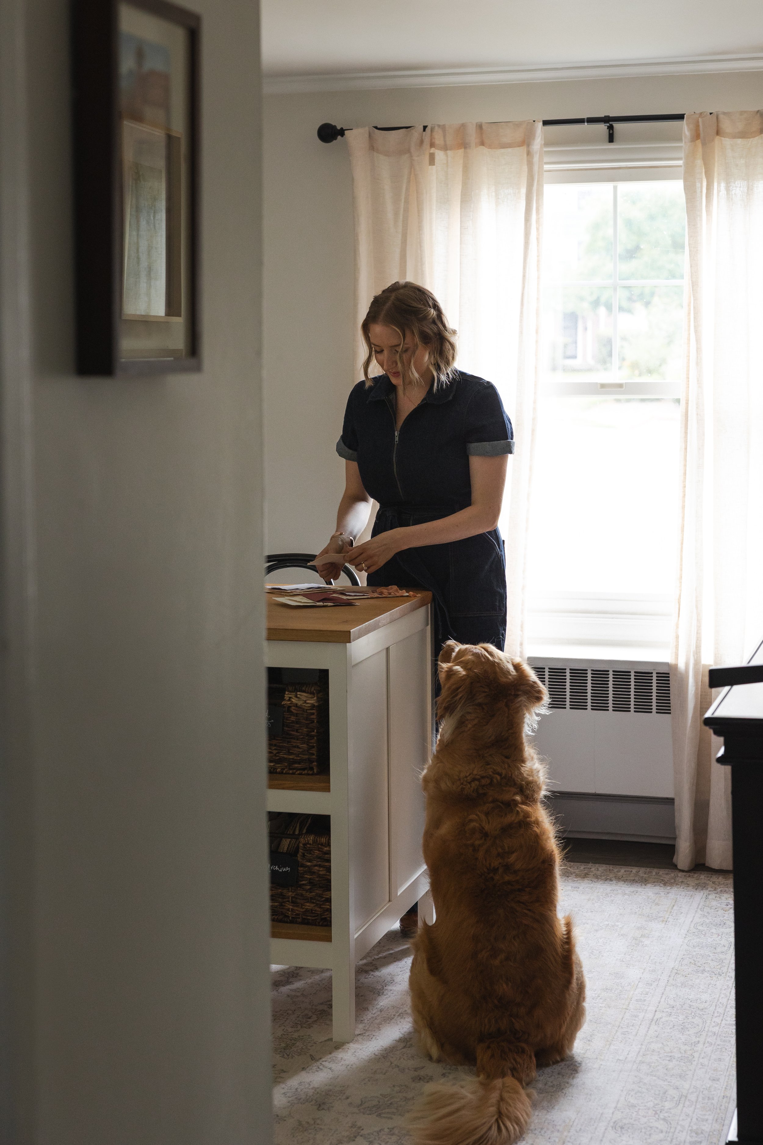 A woman with wavy blonde hair and a navy blue dress with rolled-up sleeves stands in a sunlit kitchen, looking at papers on a table. A golden retriever sits on the floor facing her, with its back to the camera. The room features cream-colored curtains, a window with a view of trees, and a white cabinet with open shelves.