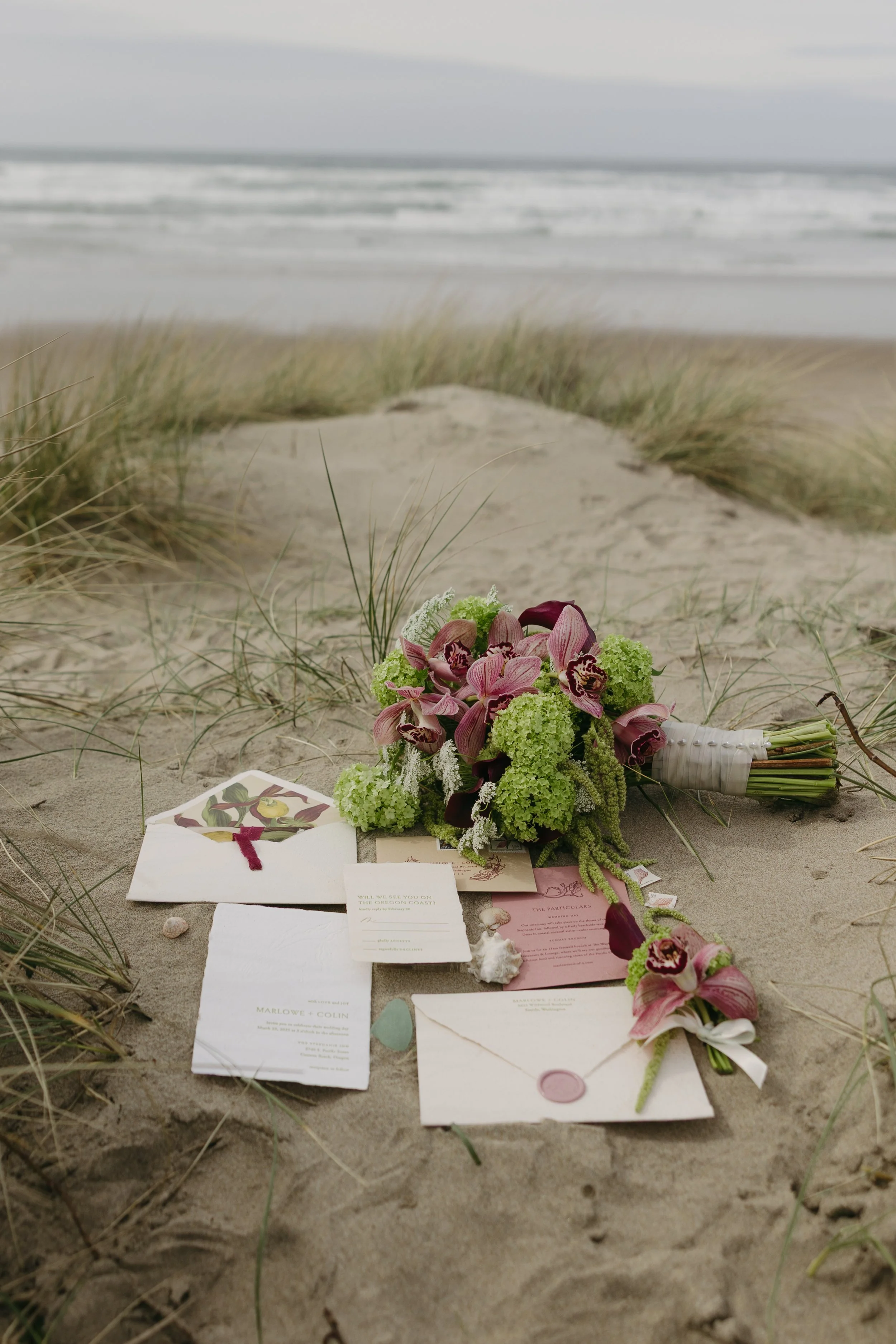 A wedding arrangement on the sand with flowers, greeting cards, and envelopes, with beach grass and ocean waves in the background.