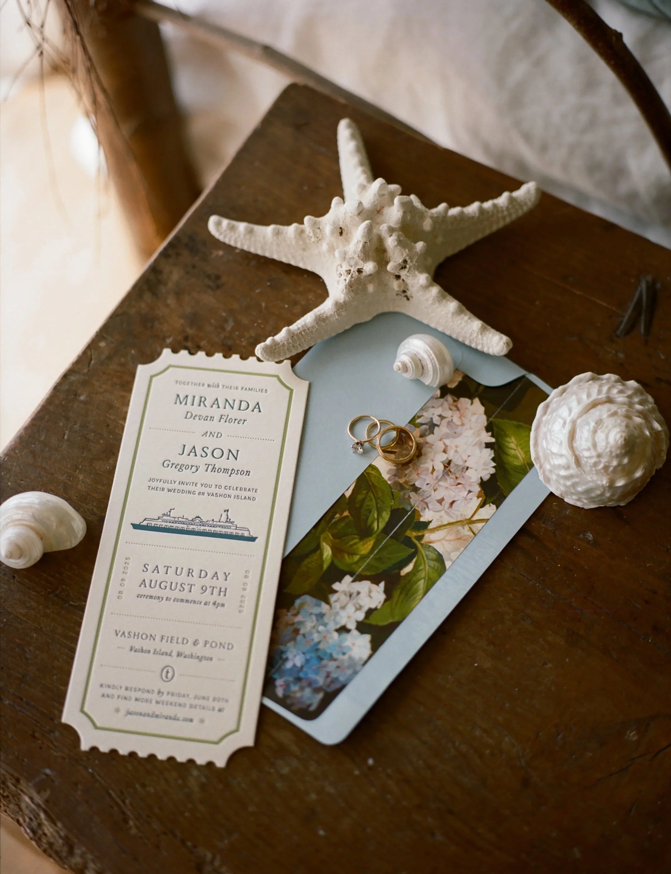 A wedding invitation card on a wooden table, decorated with seashells and a starfish, and wedding bands.