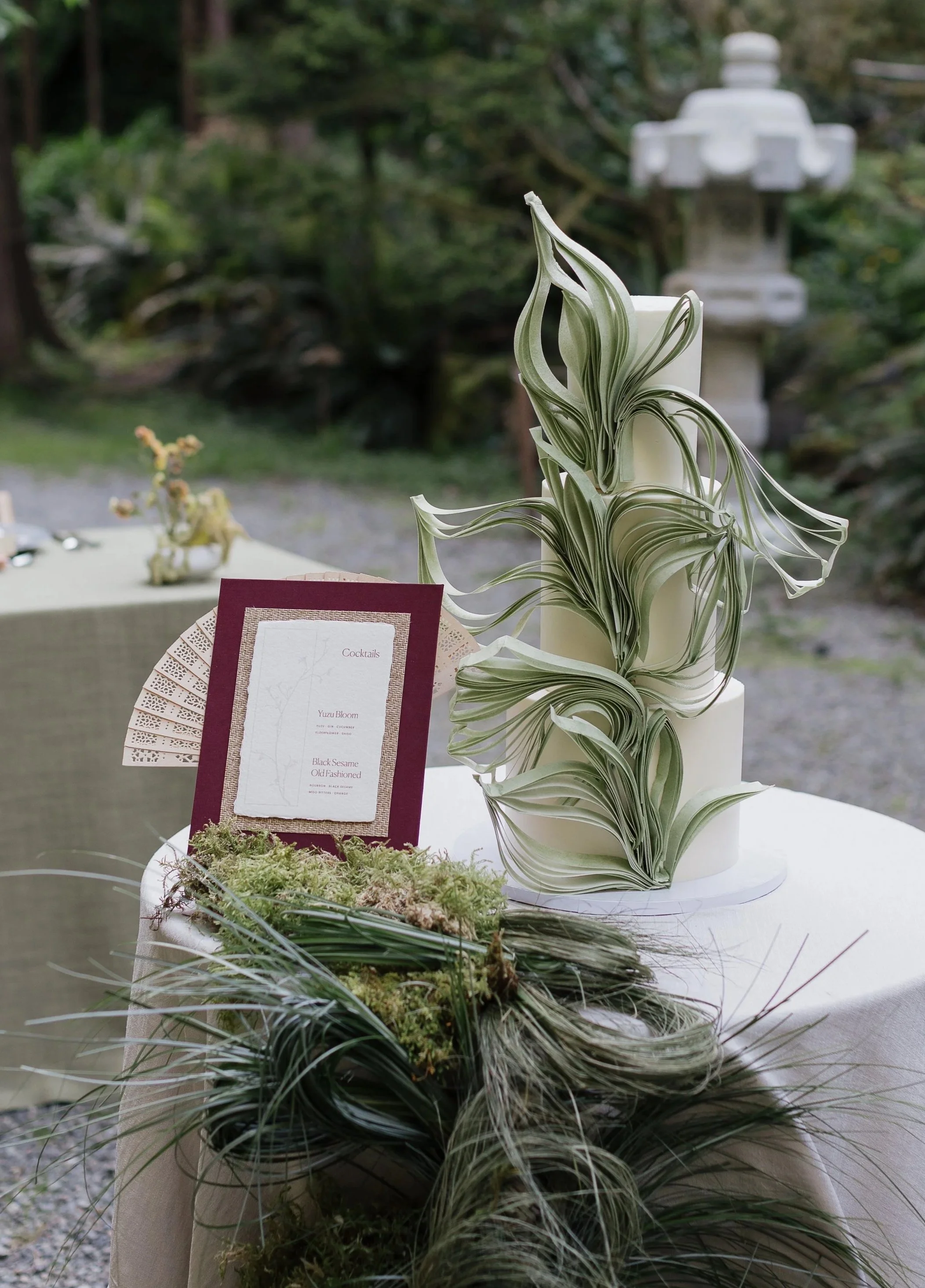 A sculptural wedding cake with intricate green paper leaves around it, placed on a table with a bar menu card and moss, outdoors in a Japanese garden.