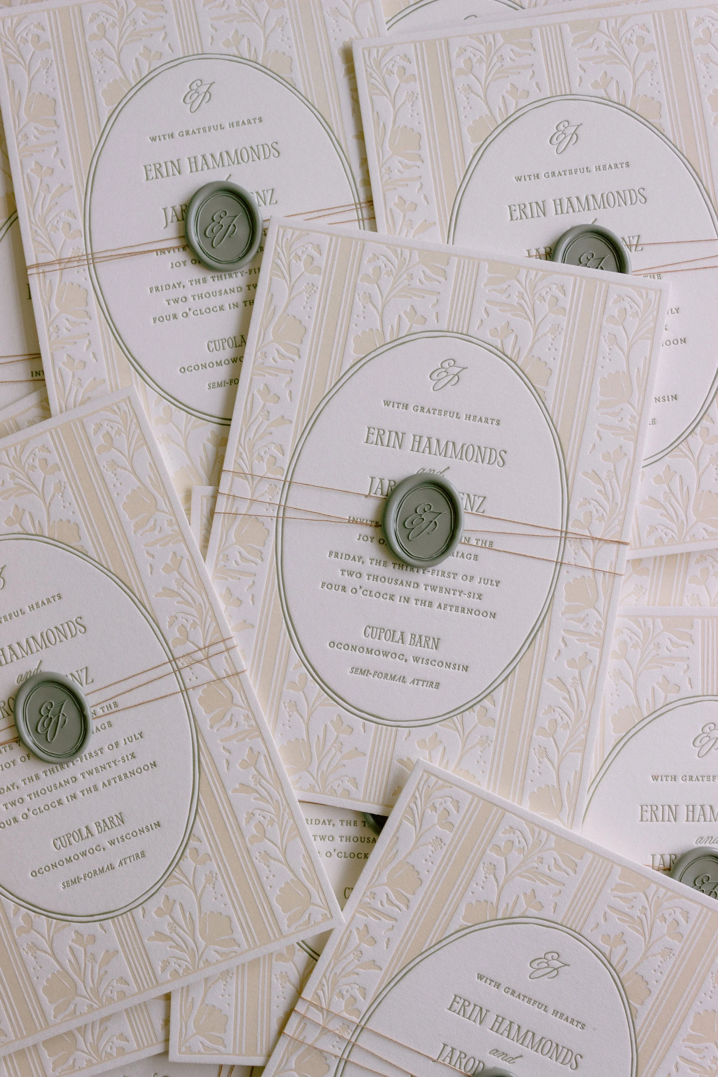 Multiple wedding invitations with a floral pattern, oval frames, and green wax seals with initials.