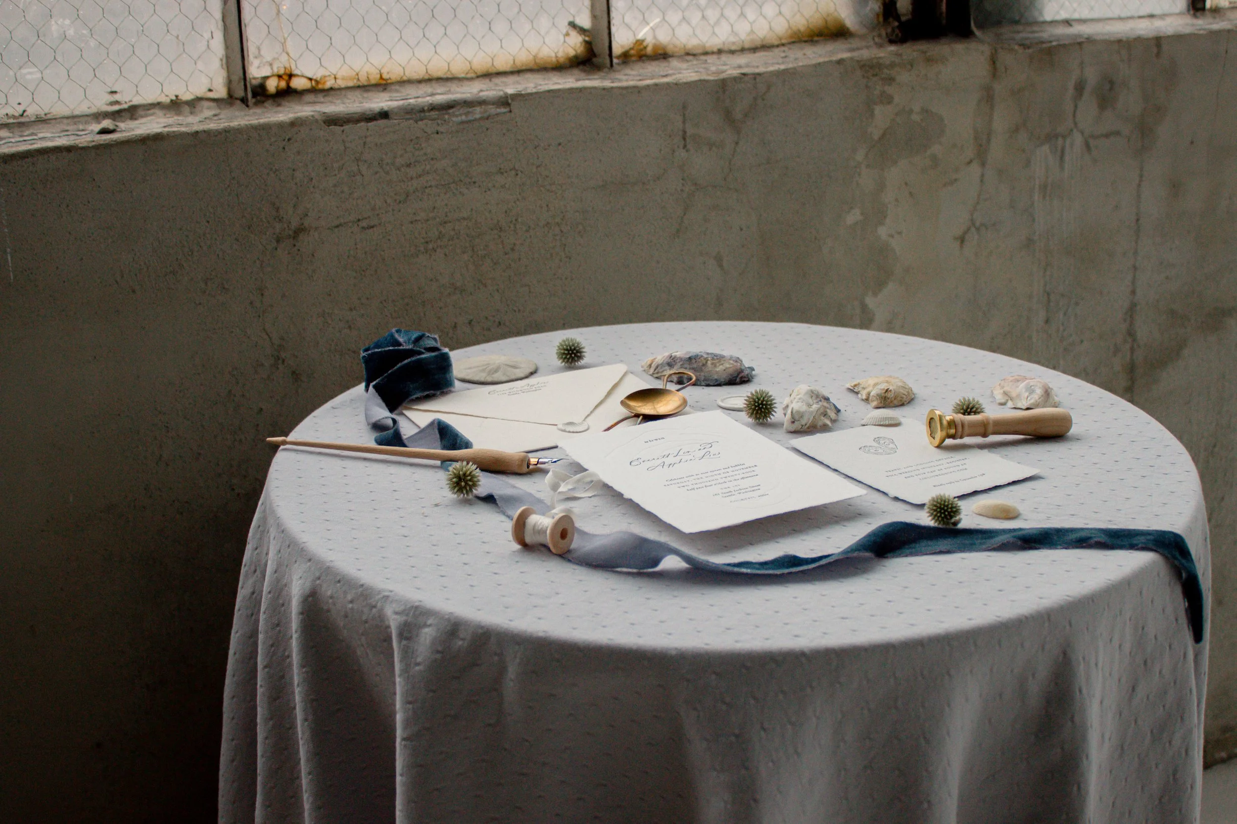 A round table with a white polka dot cloth, set against a concrete wall and chain-link fence window. The table has various objects: shells, small bulby plants, a golden spoon, an open white envelope, and some papers.