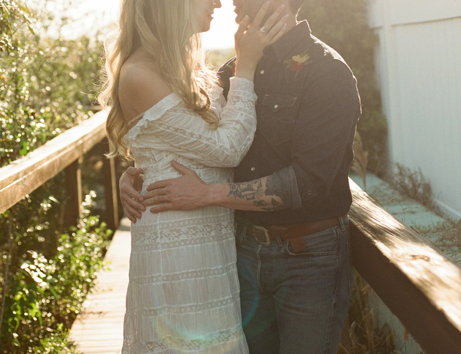 A couple embracing and about to kiss on a wooden porch during sunset, with natural greenery in the background.