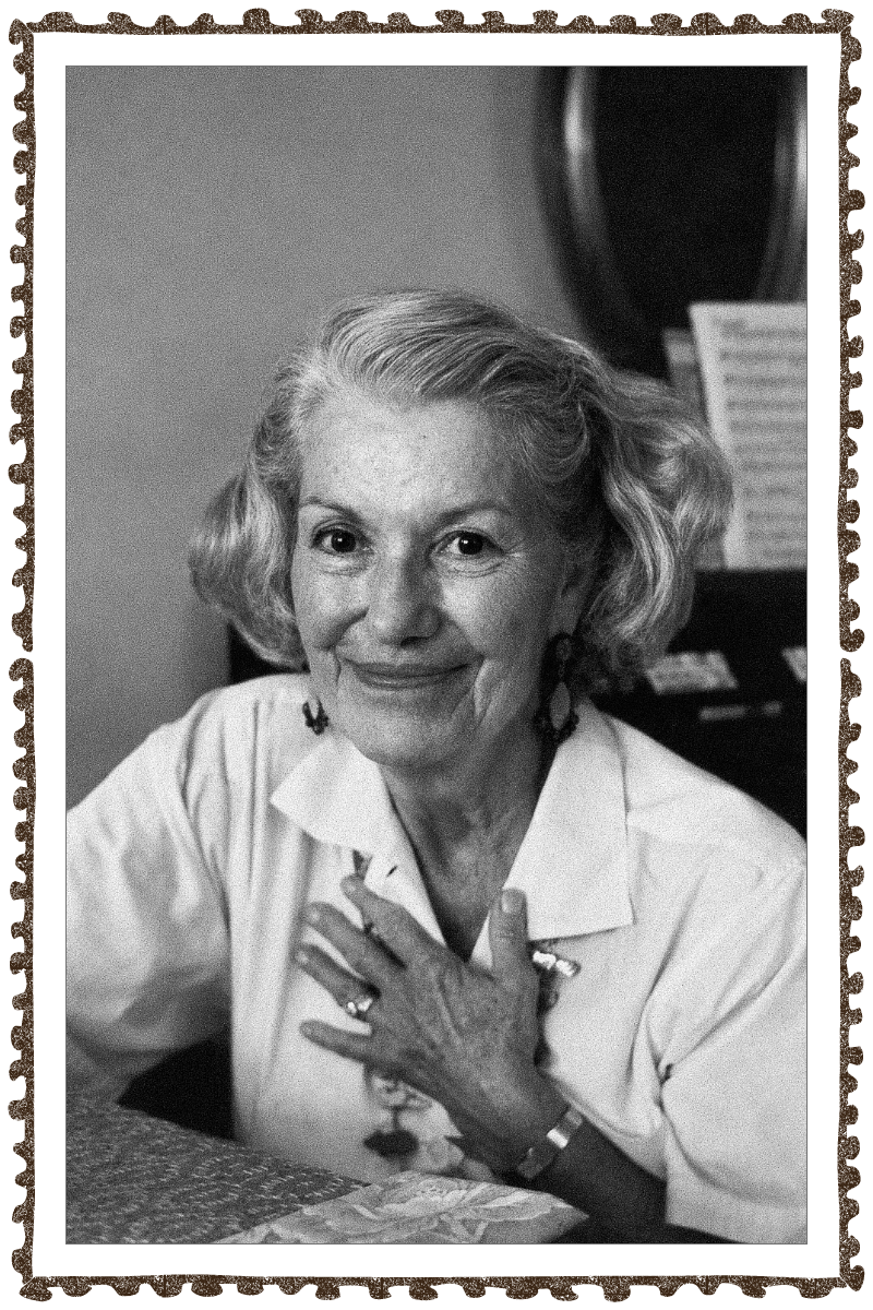 Black-and-white portrait of an elderly woman with curly hair, wearing a white shirt and jewelry, sitting at a table with an expressive background that includes a mirror and a music stand with sheet music.