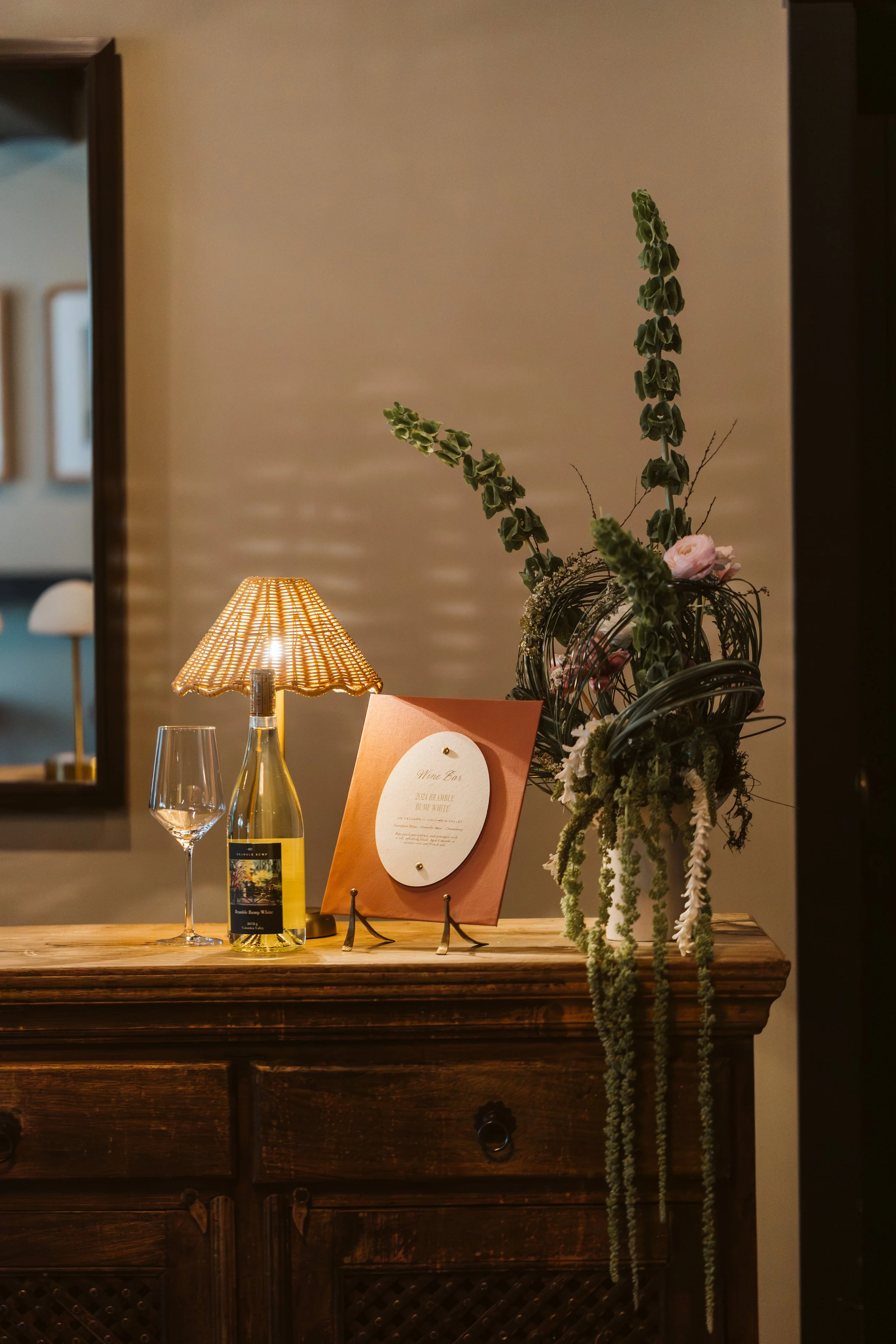 A cozy indoor setting with a wooden table, a lamp with a woven shade, a bottle of white wine, a wine glass, and a floral arrangement with tall green stems and pink flowers. There is a menu or card on a stand on the table. The background features a beige wall and a mirror.
