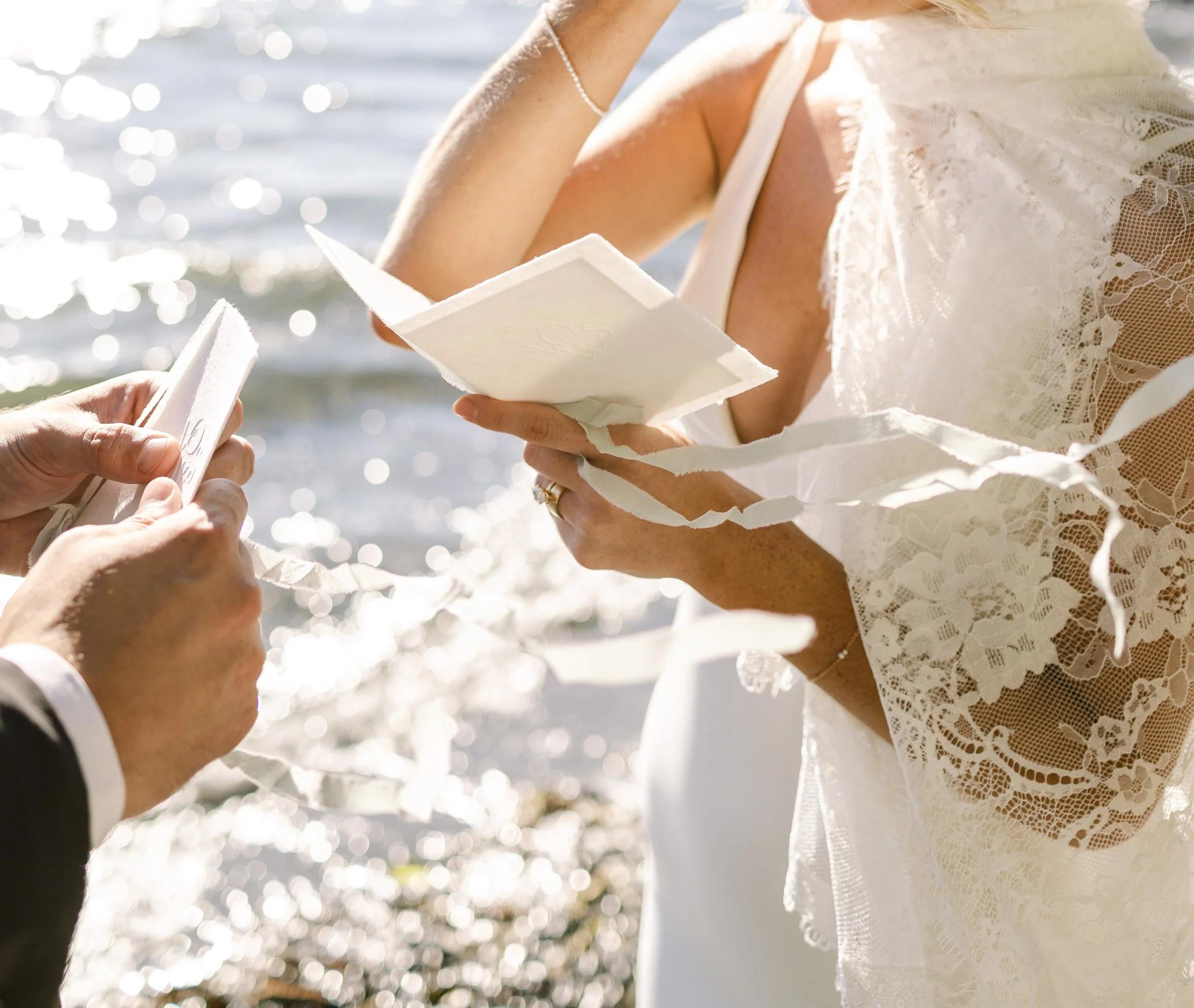Close-up of a woman in a white lace dress and a pearl necklace holding letterpress vow books during a wedding ceremony near the ocean.