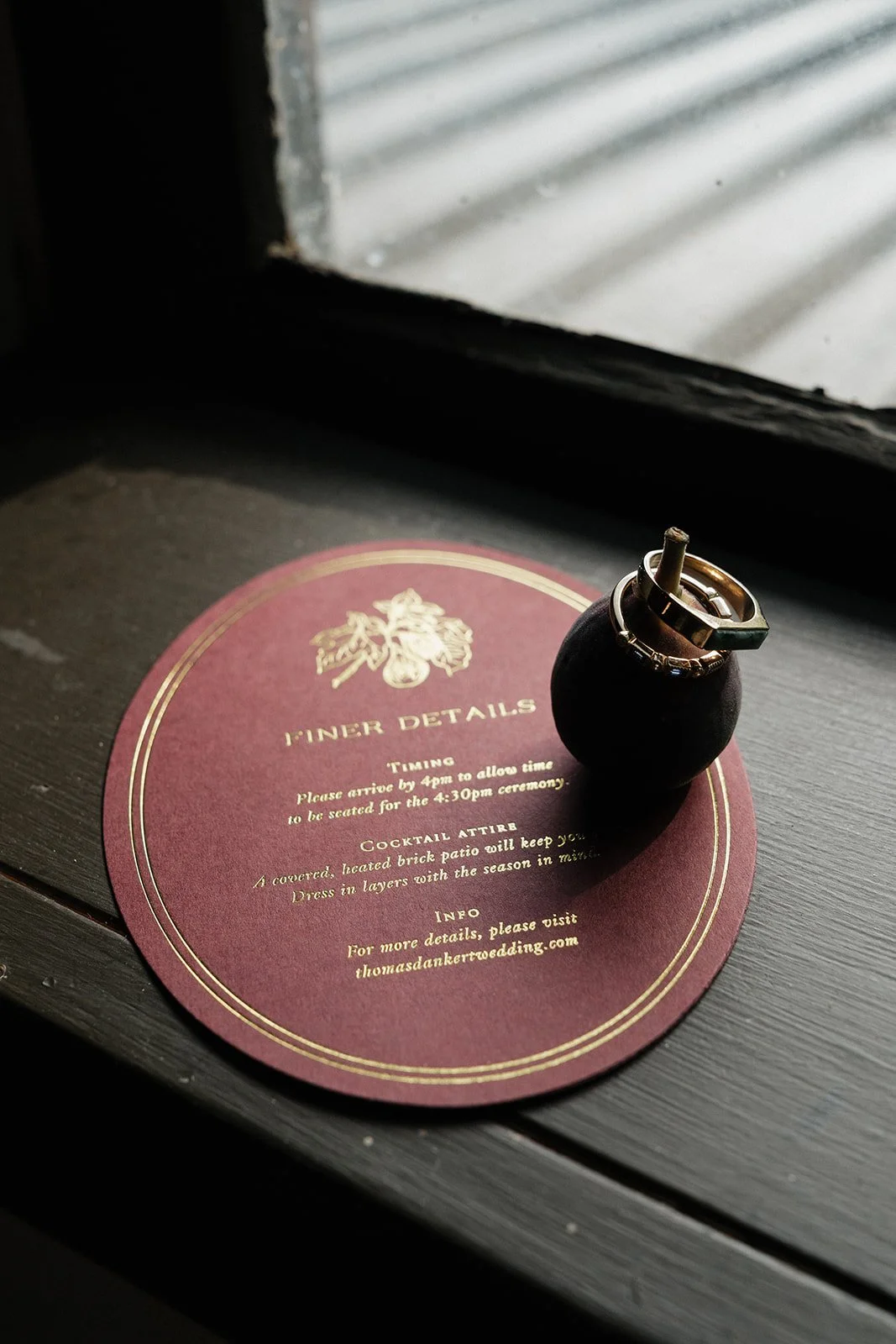 A wedding invitation card titled 'Finer Details' with gold text on a maroon background, placed on a dark table near a window. Two rings are placed on a small black ring holder on top of the card.