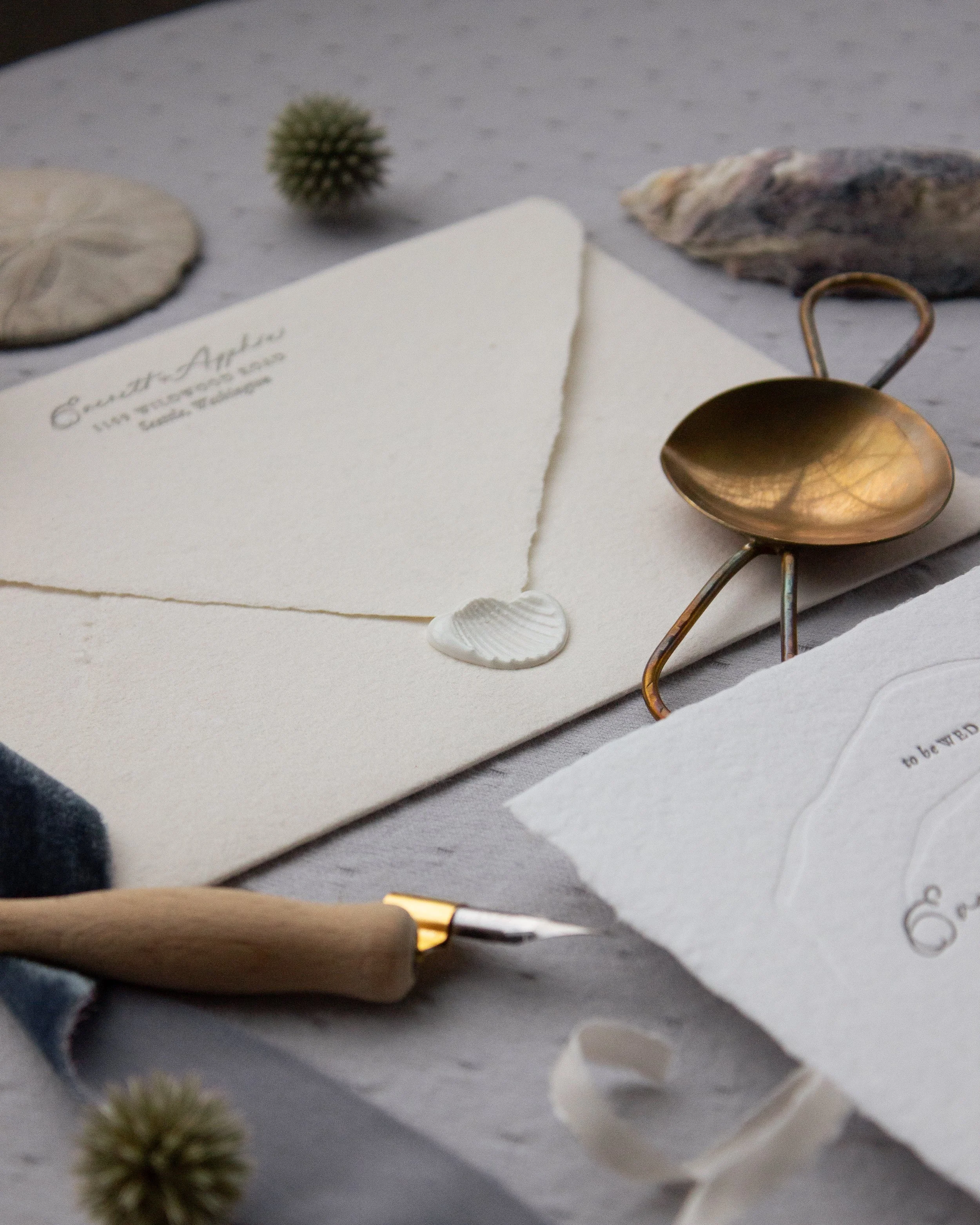 Close-up of a textured white envelope with a seashell wax seal, flanked by a small dried flower and a partially visible spoon with a metal handle on a gray surface.
