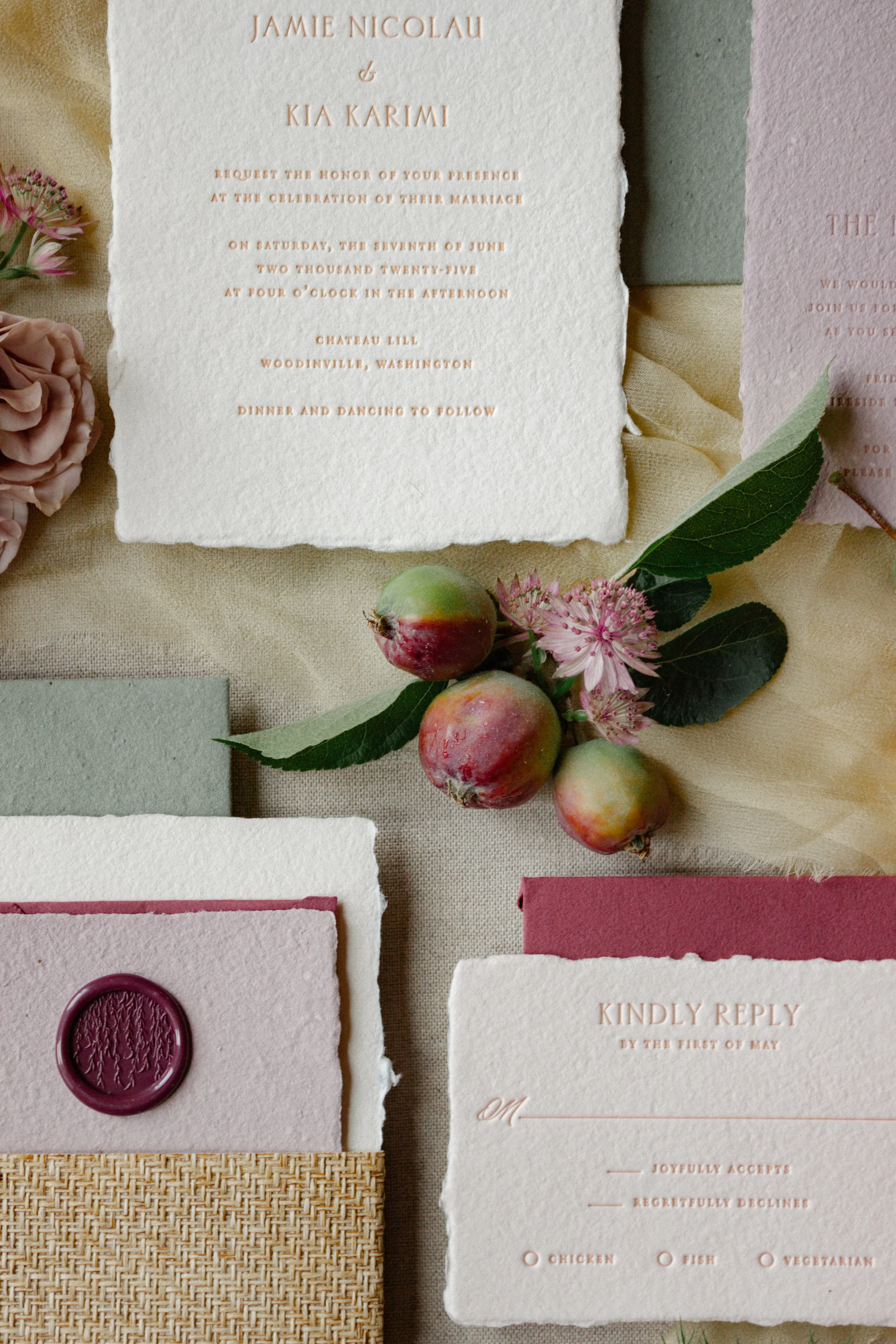 Wedding invitation card with gold text on textured paper, surrounded by small pink and green decorative elements, including flowers, a wax seal, and fruit.