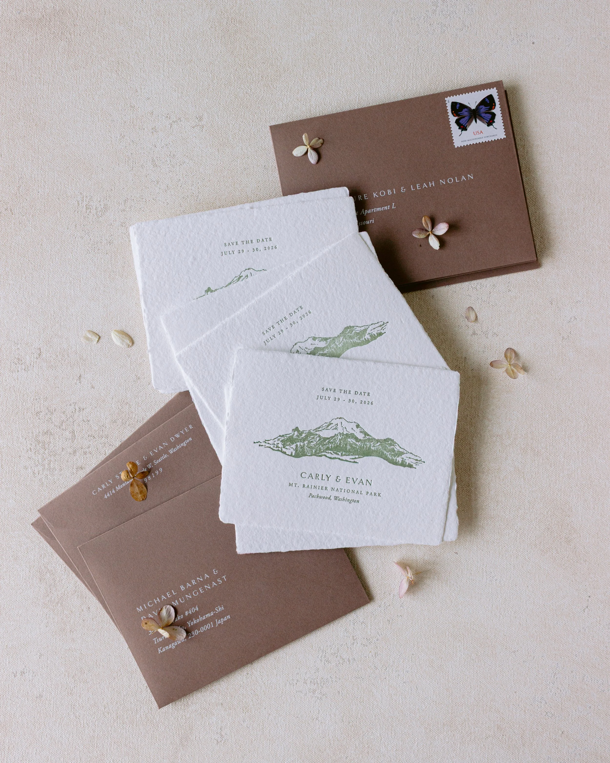A collection of wedding or event invitations and cards laid out on a light-colored fabric surface. The invitations include details about the event such as dates, locations, and names, with some featuring mountain illustrations. Small flower-shaped decoration and a few flower petals are scattered around.