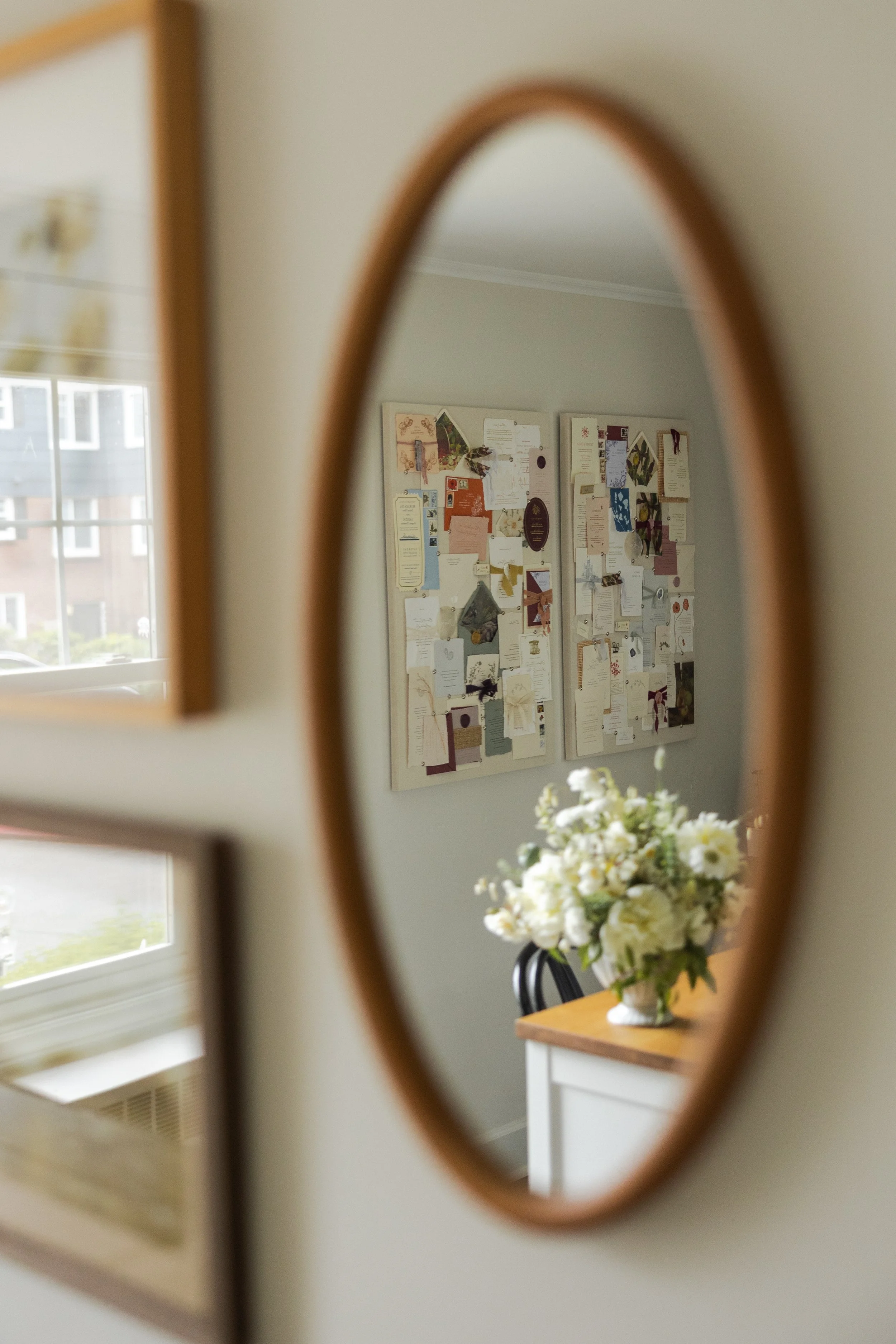 A reflection in an oval mirror shows a wall with two bulletin boards filled with various papers and decorations, a window with visible buildings outside, a table with a white vase filled with white flowers, and a chair.