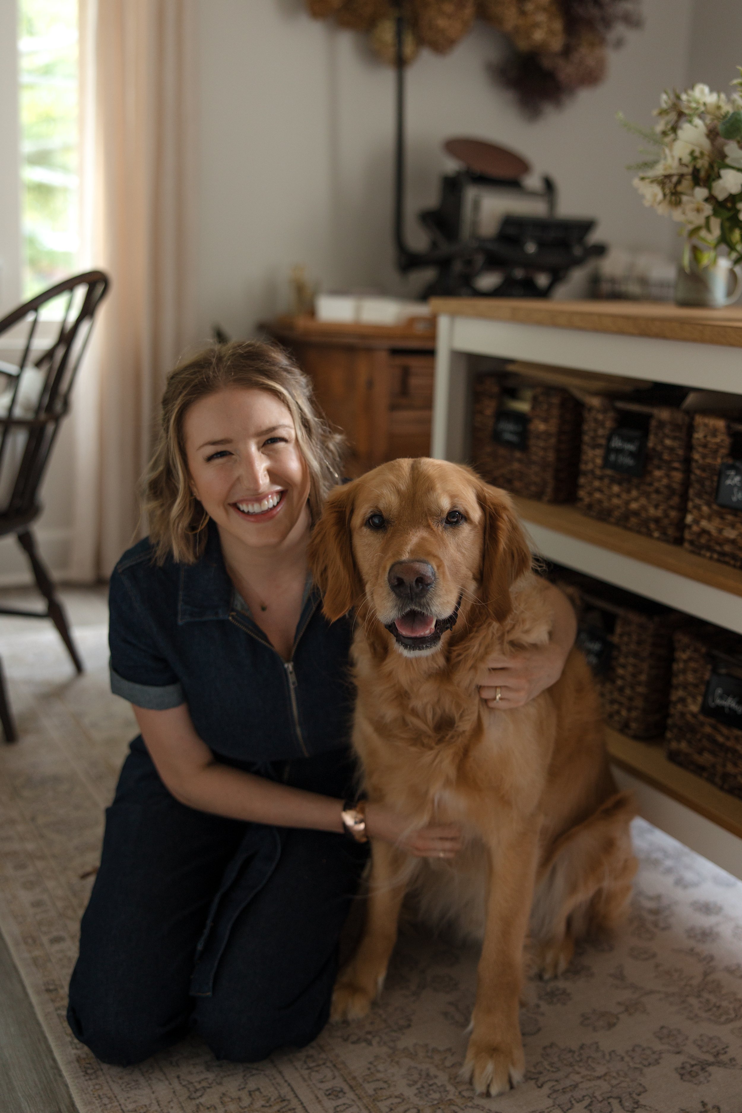 A woman smiling and kneeling on a patterned rug with a large golden retriever dog indoors near a wooden shelf with baskets.