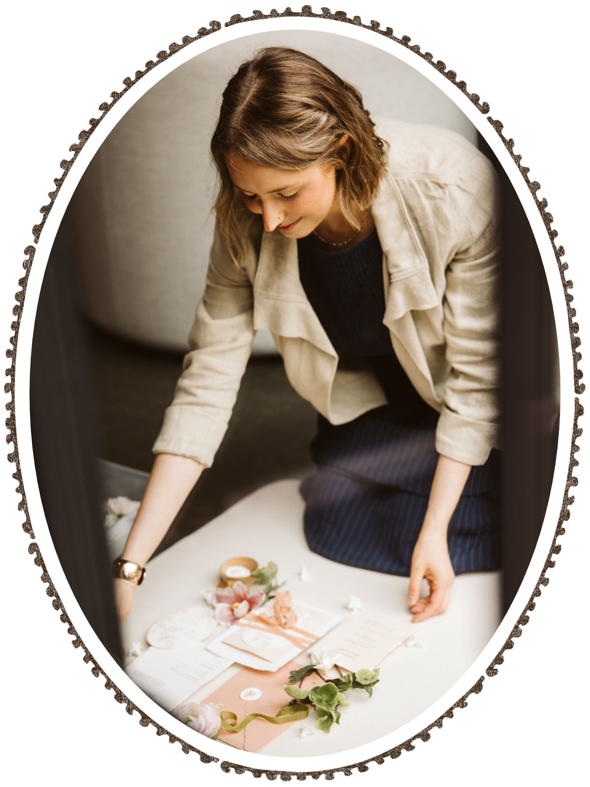 A woman in a beige blazer and black dress kneels on a table, arranging floral decorations and paper envelopes, with a cozy sofa in the background.