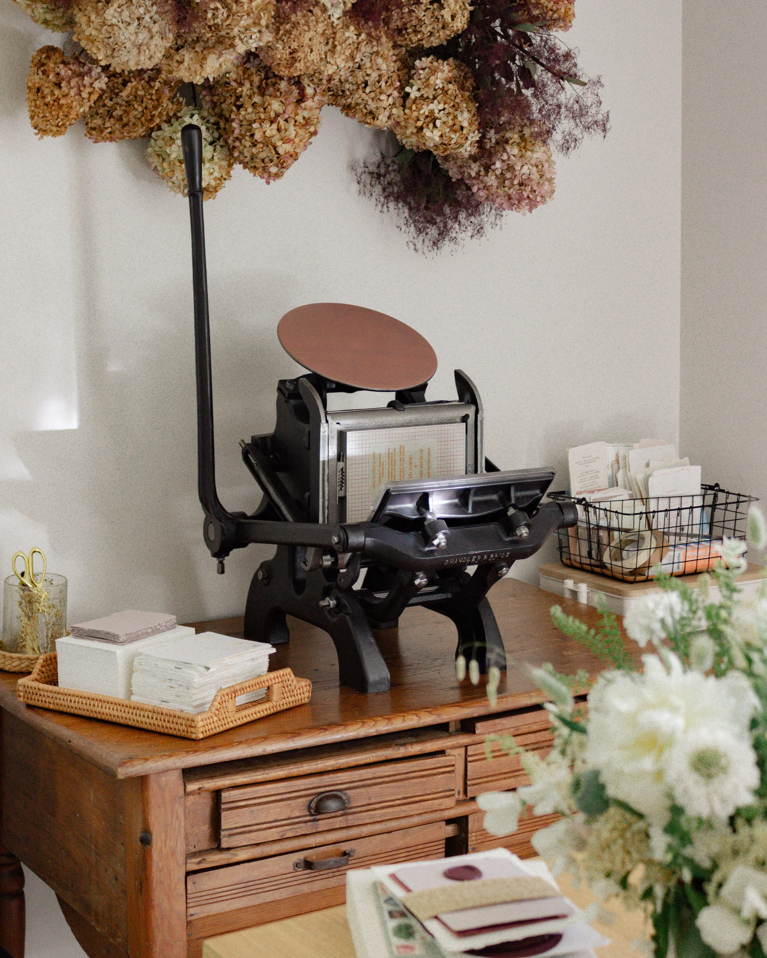 A vintage wooden drawer unit on a table with various items on top, including a small basket with napkins, a black basket with papers, and a bouquet of white flowers. In the background, there is a black file organizer with papers and an orange ruler, and a lamp with a black adjustable arm shining light on the objects.