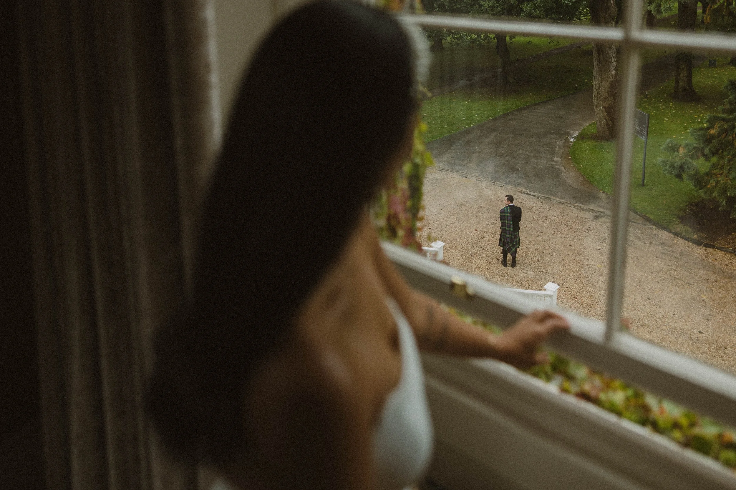 A girl looking out a window at a person walking outside on a gravel path in a park-like setting during rainy weather.