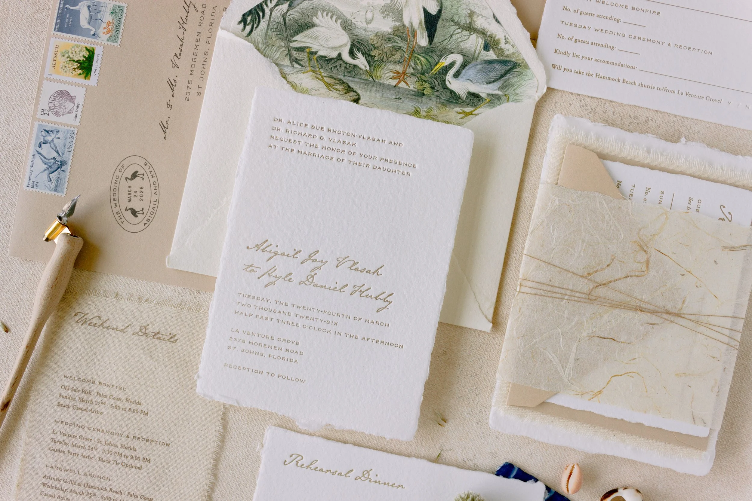 Close-up of wedding invitation and details, with postage stamps, dried flowers, and decorative elements on a beige table.