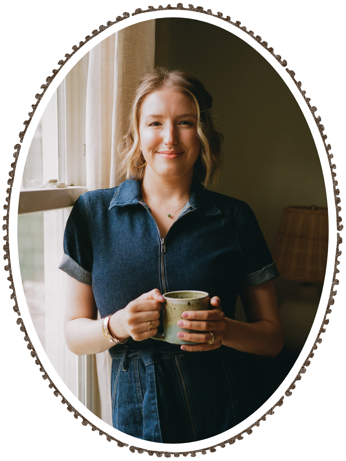 A young woman with wavy blonde hair holding a mug, standing by a window with curtains, smiling at the camera.