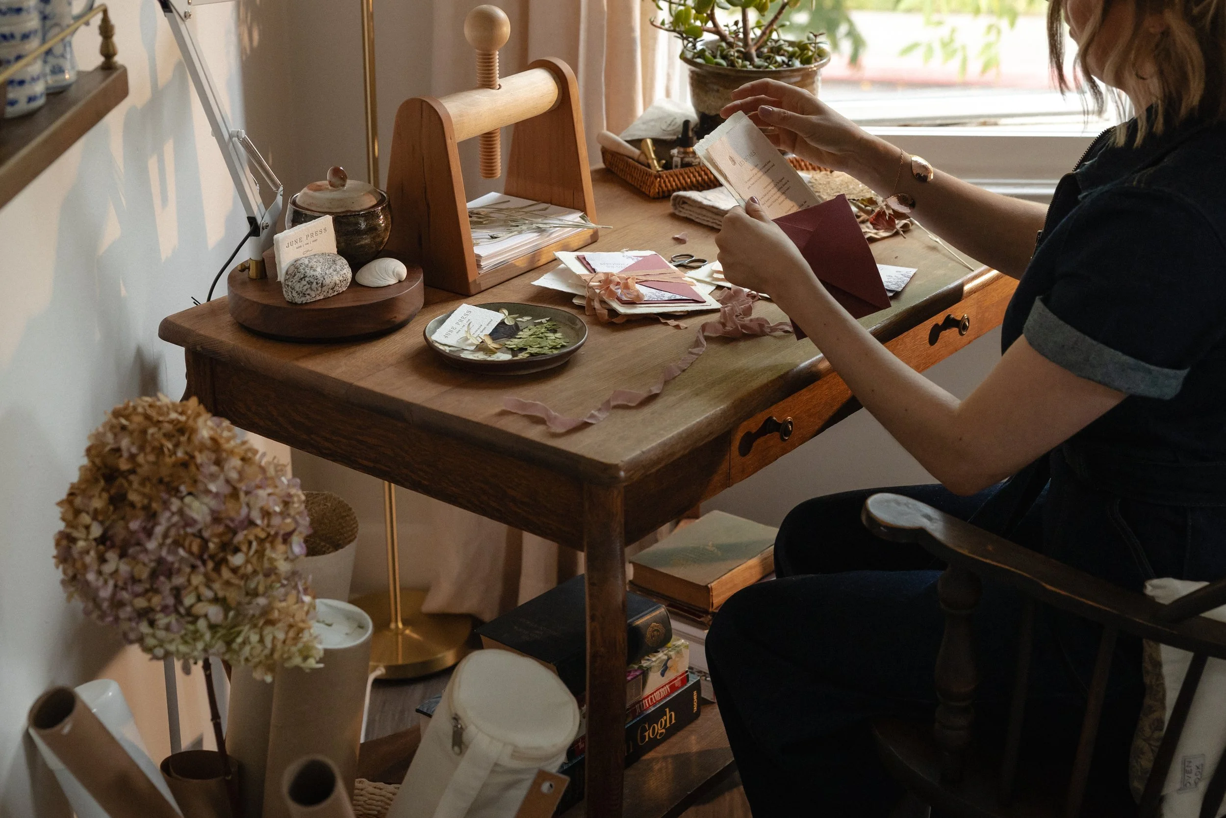 Person sitting at a wooden desk, reading a card or note, surrounded by stationery, paper, and decorative items in a cozy, well-lit room with a window and plants.