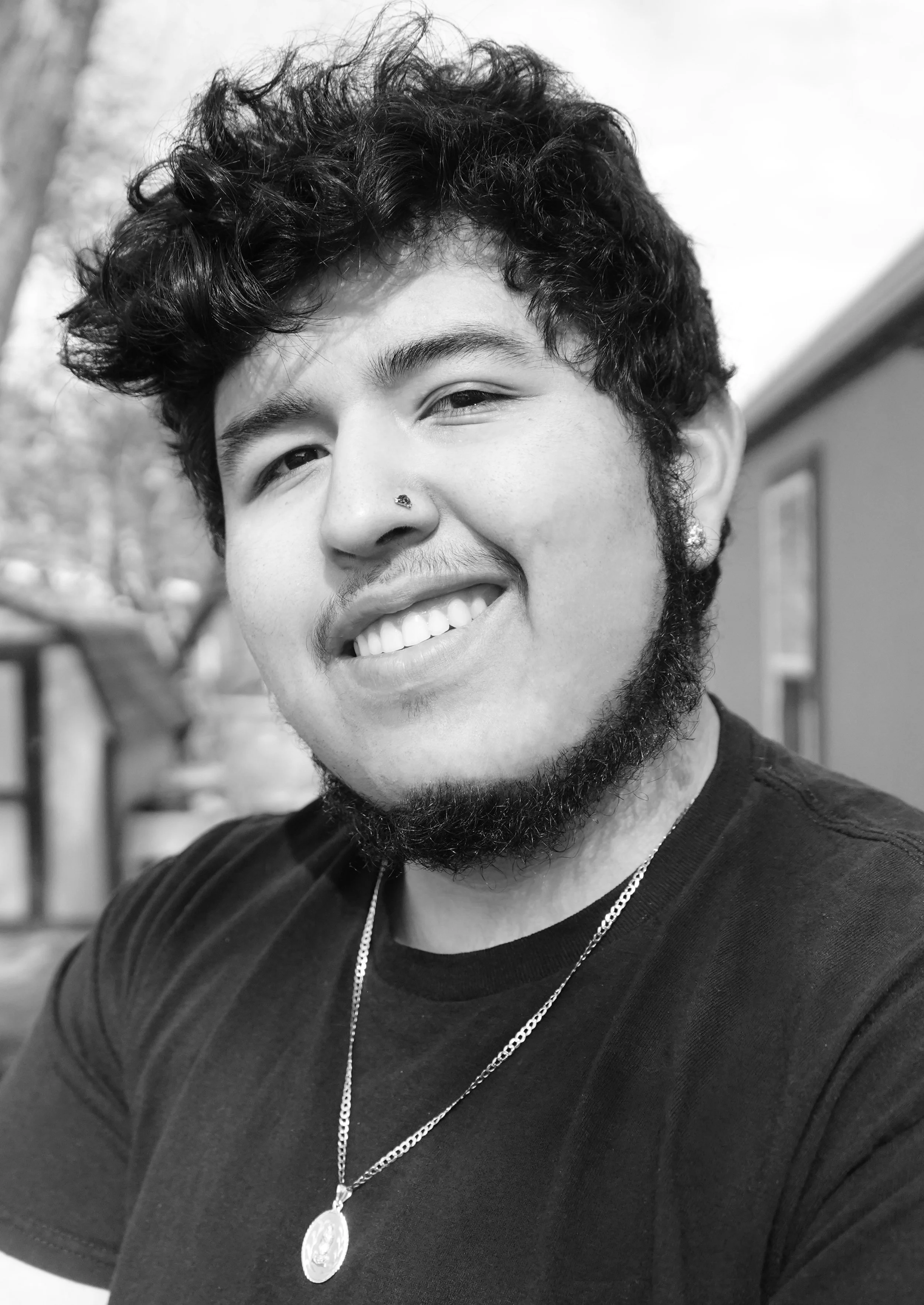 A young man with curly hair, a nose piercing, and facial hair smiling outdoors in black and white.