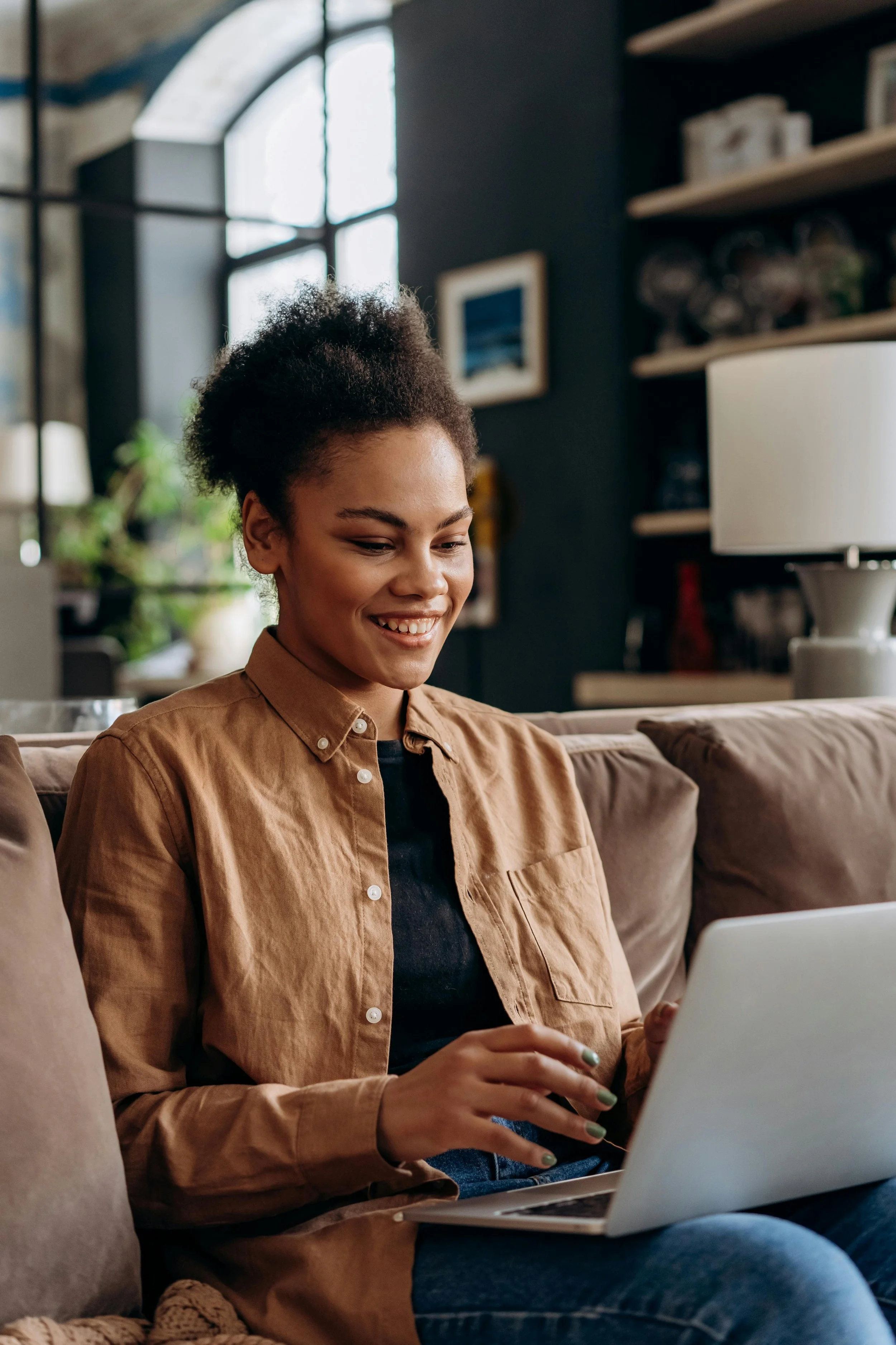 A smiling young woman sitting on a beige couch, looking at her open laptop in a cozy living room.