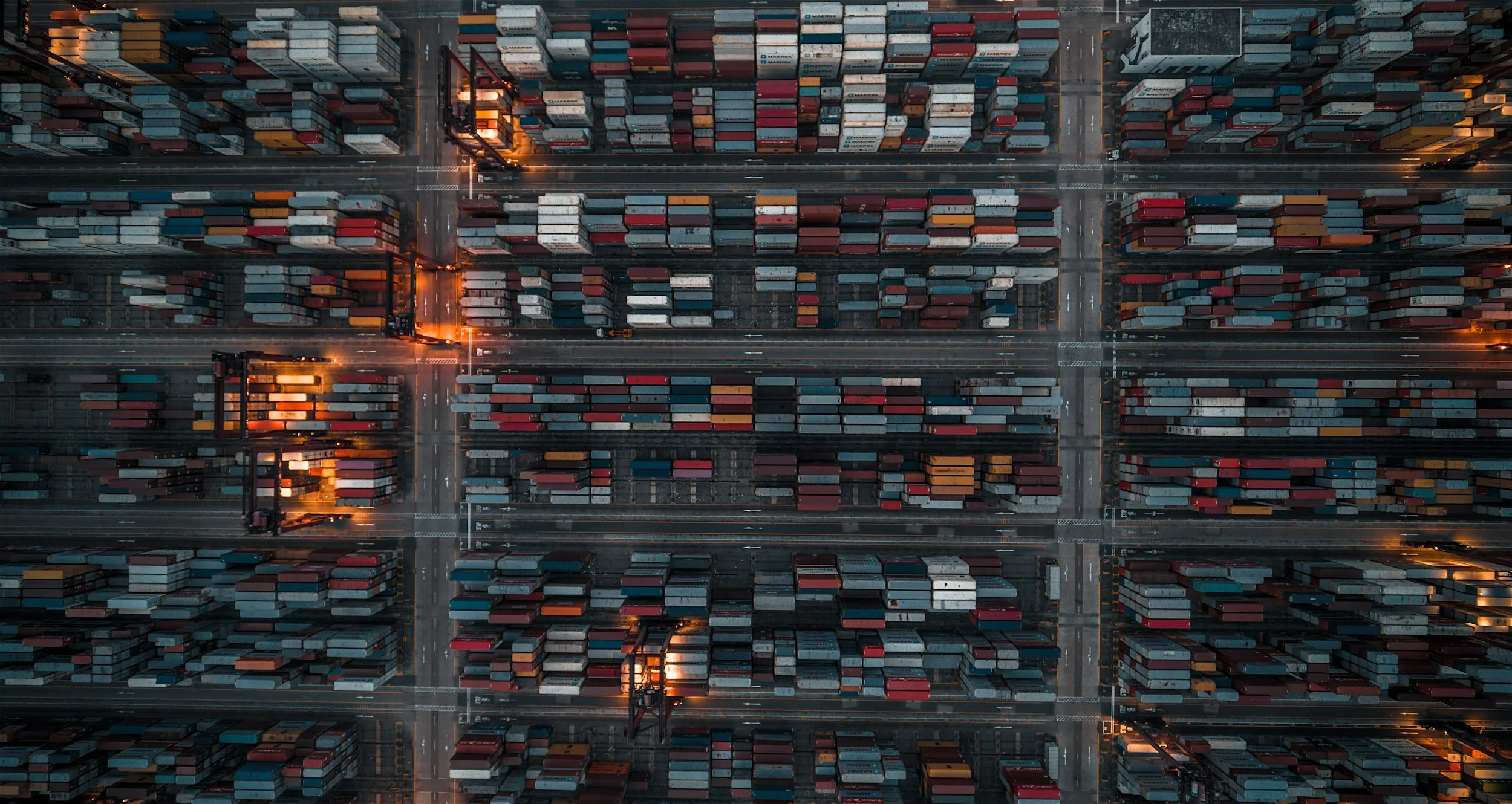 An aerial view of a shipping yard filled with numerous stacked containers and cranes, taken at dusk or early evening.