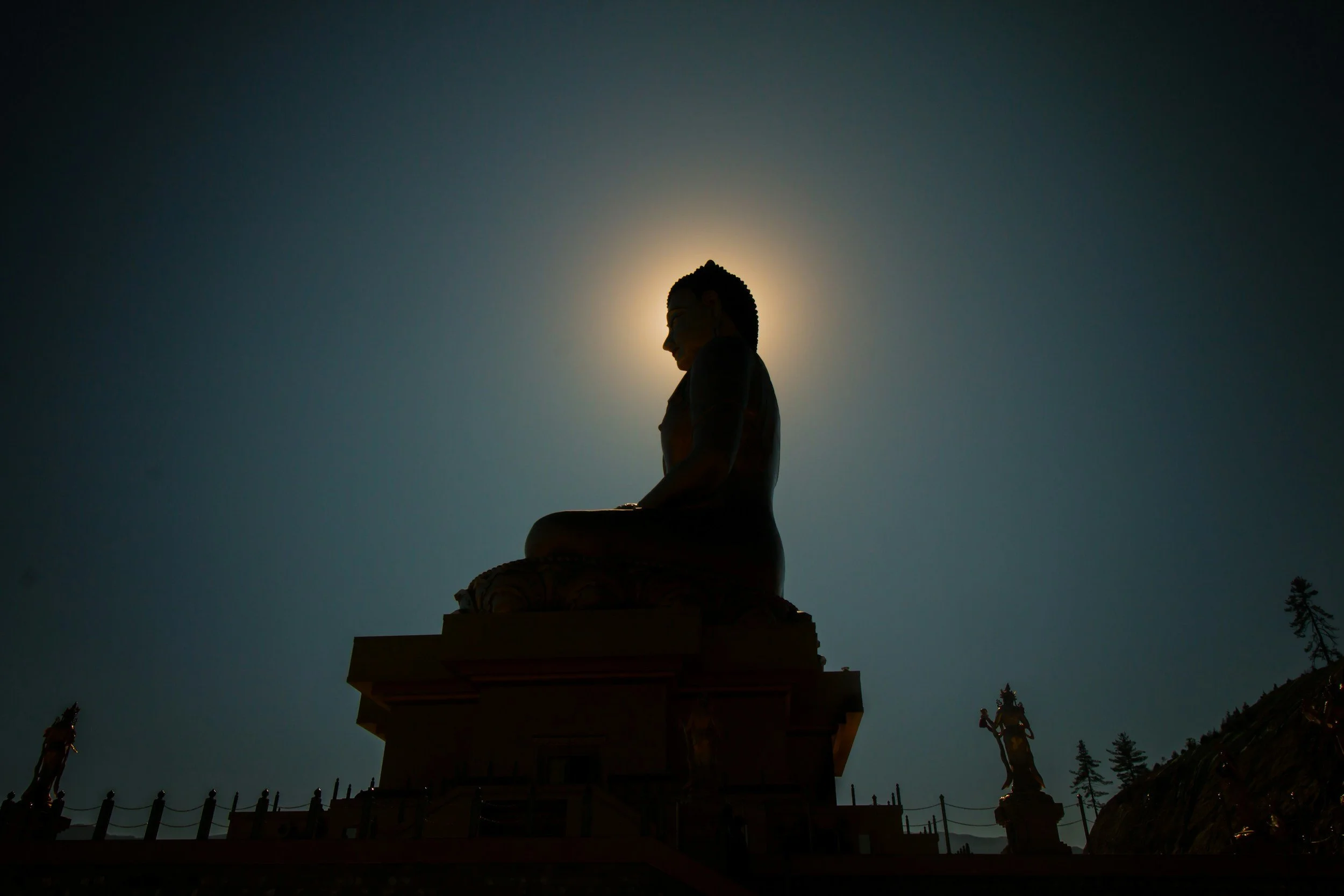 Silhouette of a seated Buddha statue with the sun behind it creating a halo effect, with smaller statues and trees in the background.