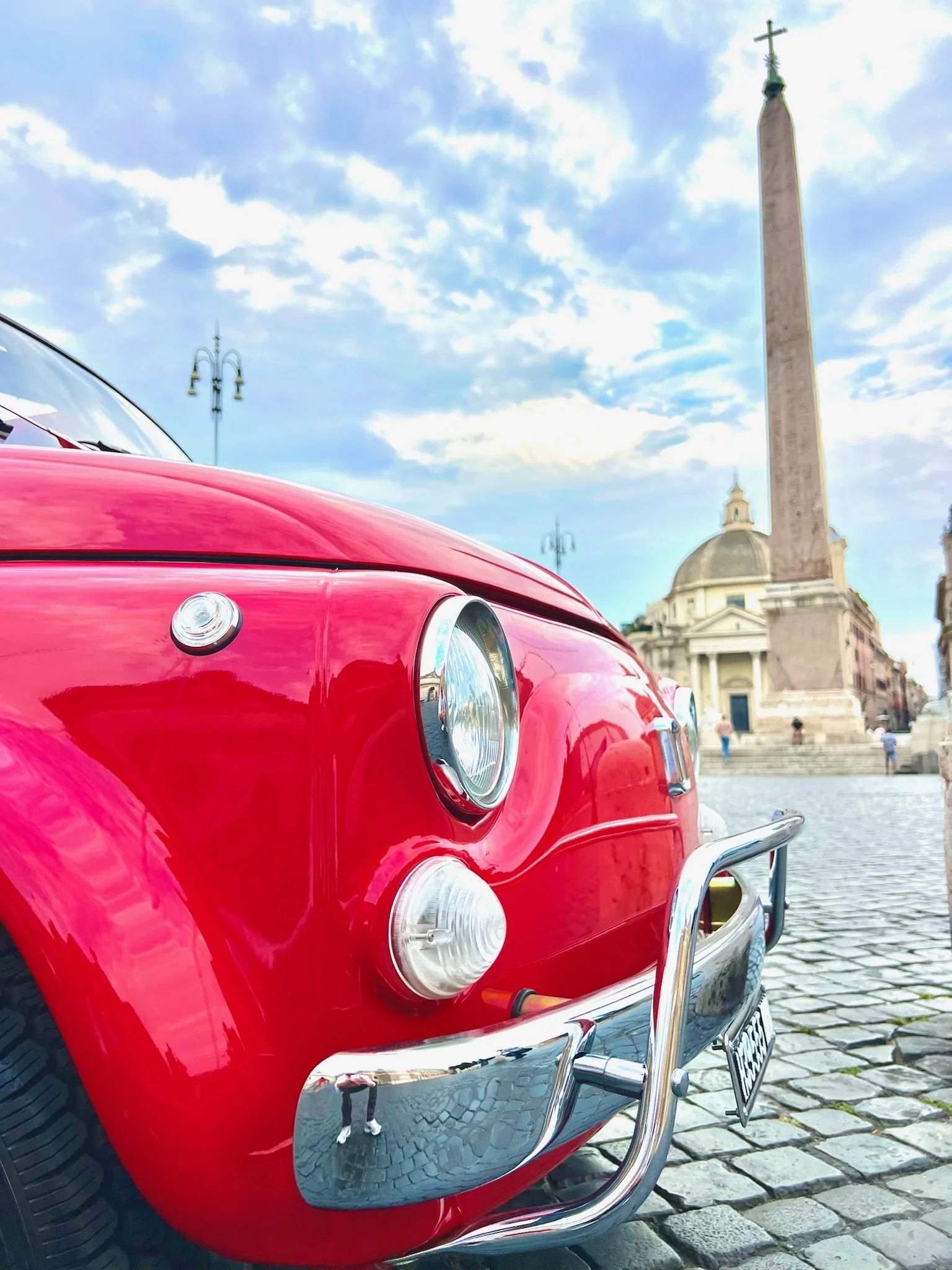 Auto rosso d'epoca davanti a un obelisco e a un edificio storico in una piazza di pietra, con il cielo nuvoloso.