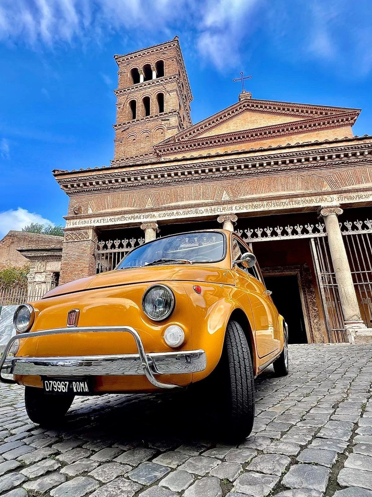 Un'auto d'epoca gialla parcheggiata davanti a una chiesa in mattoni con torretta, sotto un cielo blu con alcune nuvole.