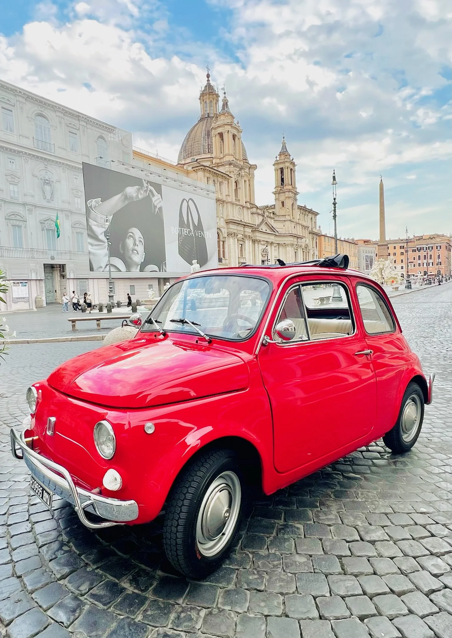 Auto rossa vintage su ciottoli in una piazza, con chiese storiche e pubblicità in background.