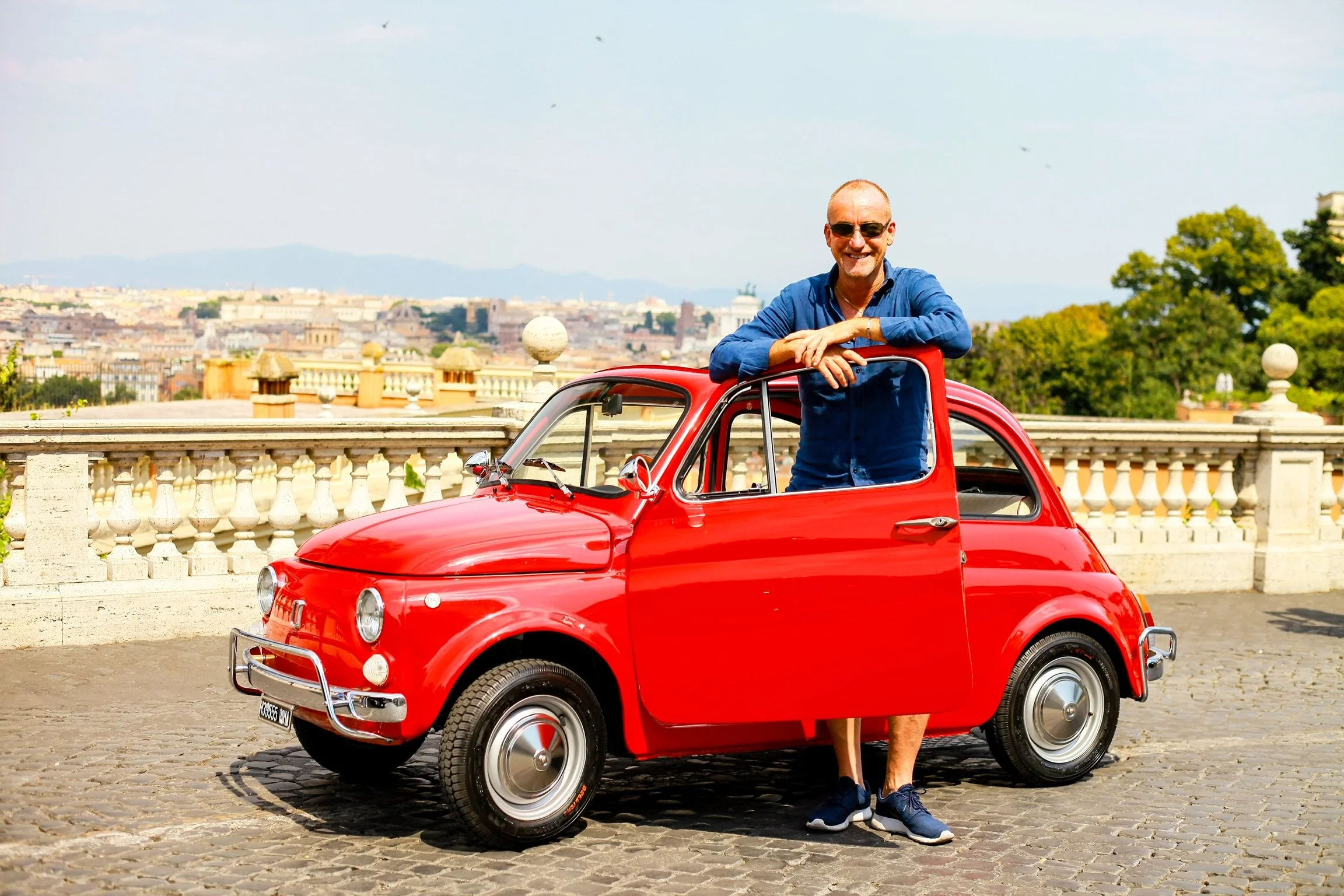 Uomo sorridente con occhiali da sole, vestito in camicia blu, che si appoggia su una piccola auto rossa, in un luogo aperto con una vista di una città e natura sullo sfondo.