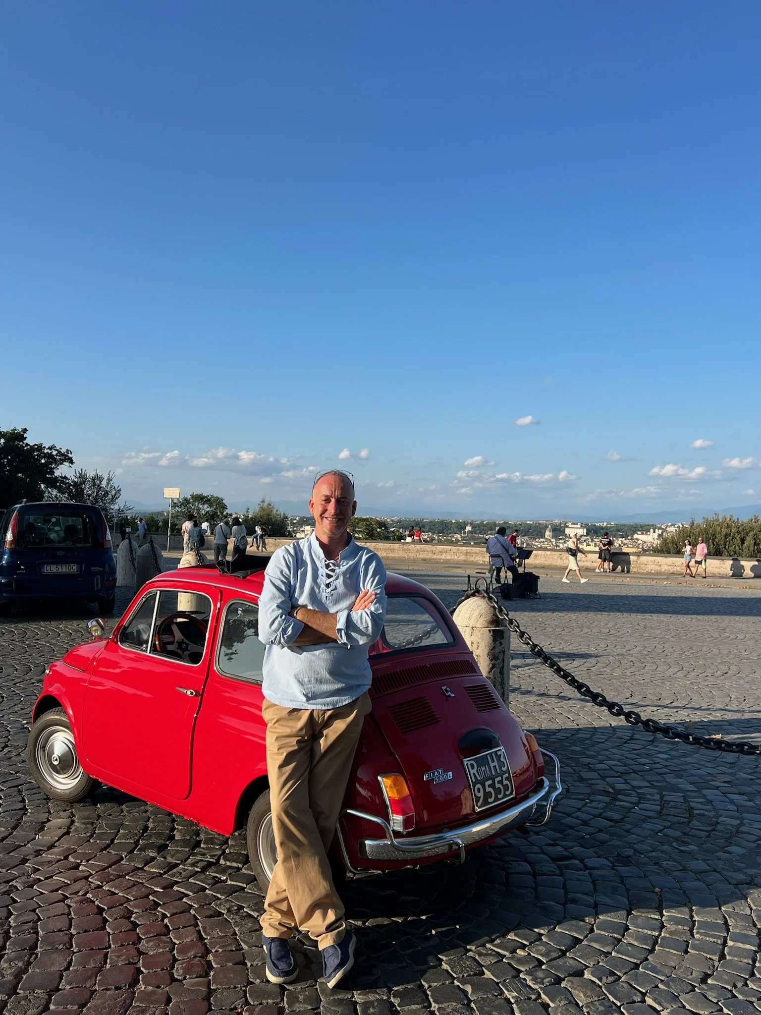 Uomo felice che si trova davanti a una macchina rossa Fiat 500, con sfondo di una passeggiata in strada e persone che camminano, cielo sereno e panorama urbano in lontananza.
