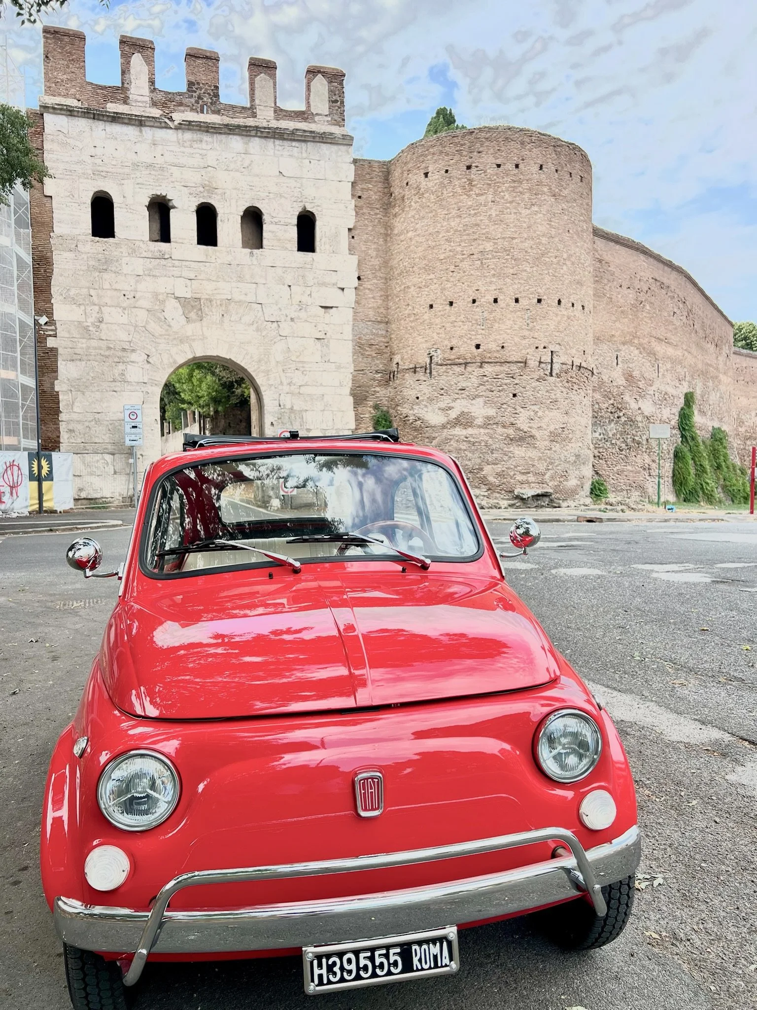 Auto rossa Fiat davanti a una vecchia porta di città con mura diroccate, sotto un cielo nuvoloso.