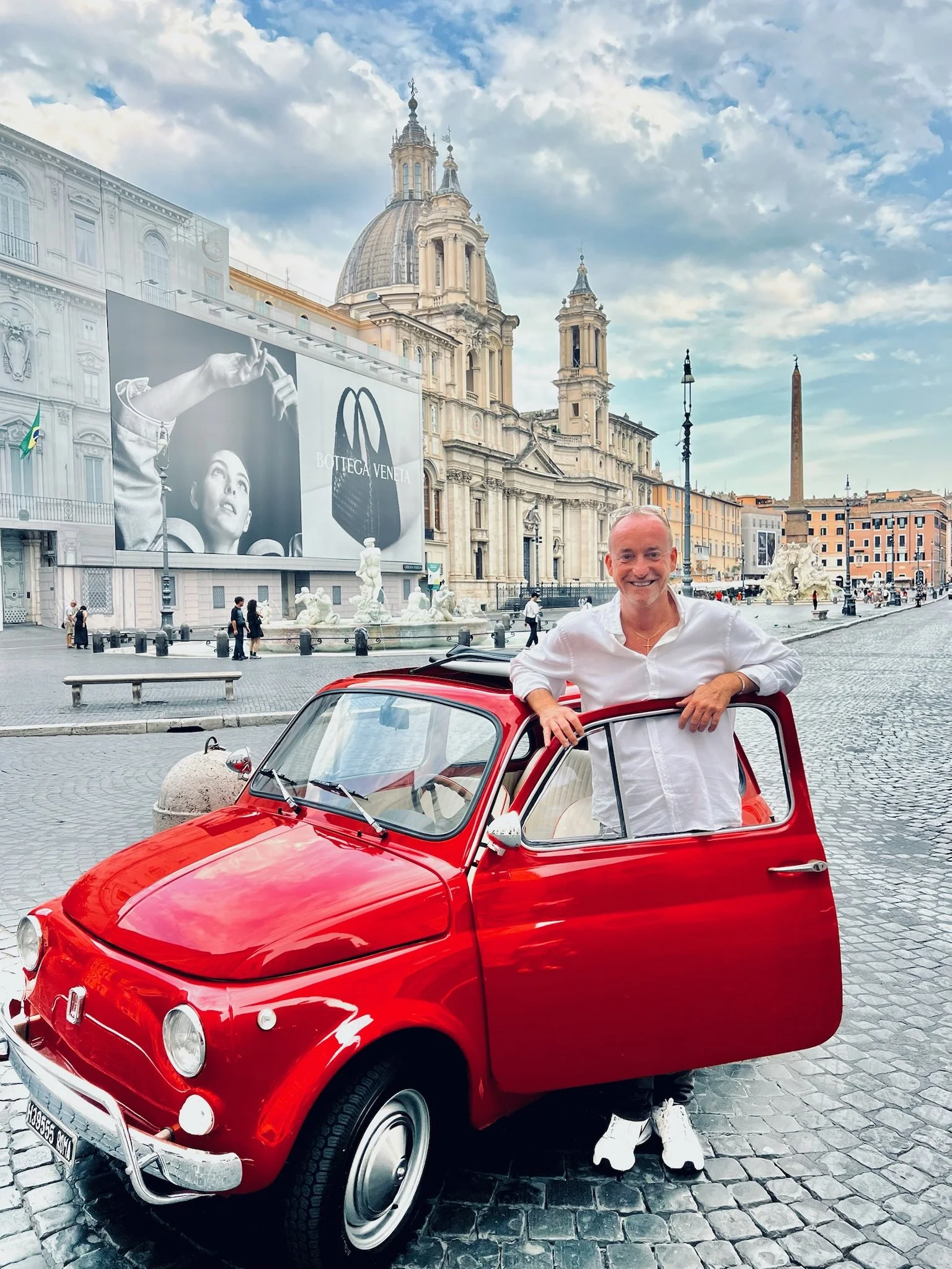 Uomo sorridente con maglietta bianca e scarpe da ginnastica bianche, che si appoggia alla portiera aperta di una piccola auto rossa in piazza con chiesa storica sullo sfondo.