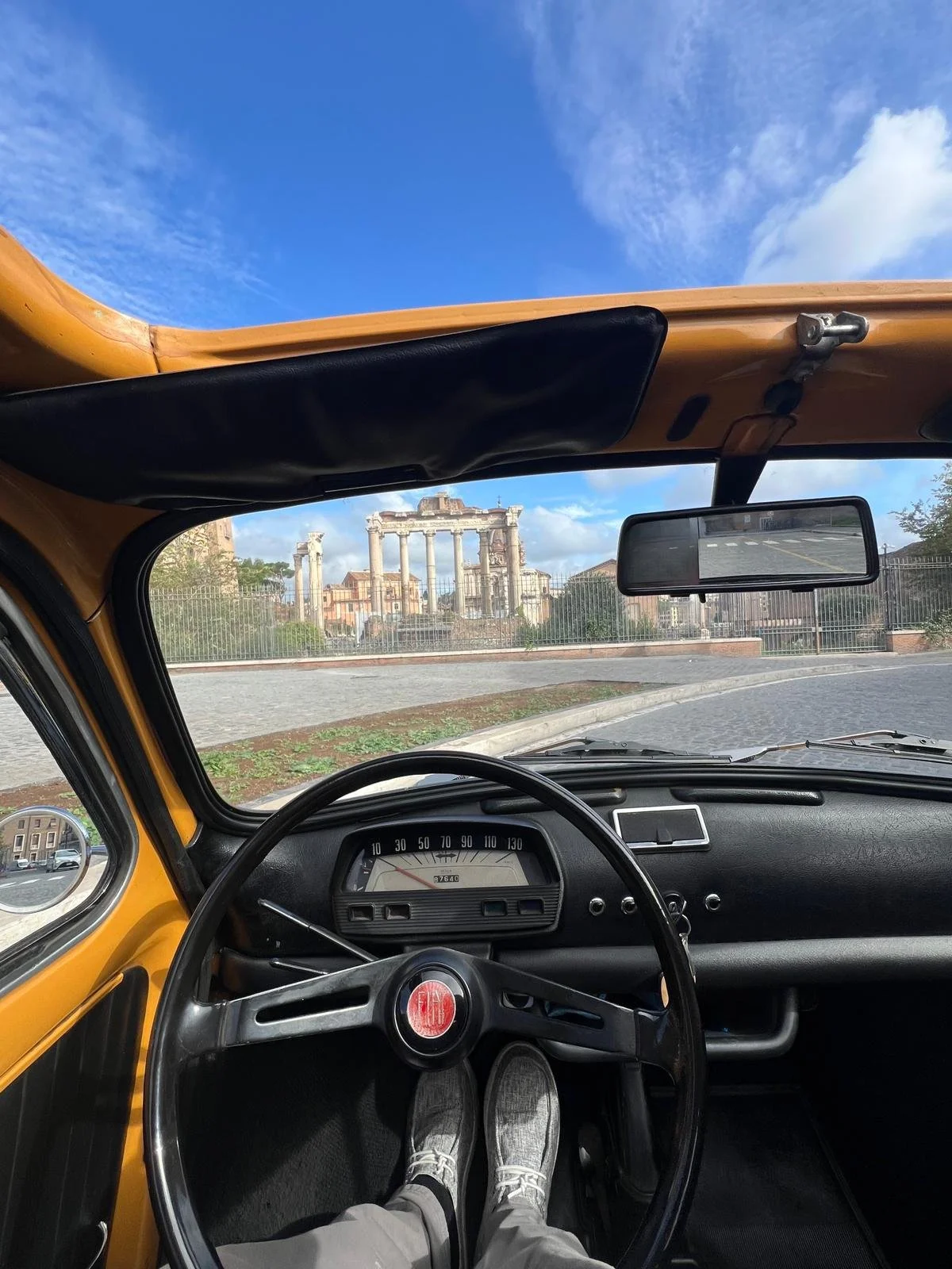 Vista dall'interno di un'auto d'epoca con il volante e il cruscotto, mostrando le scarpe del guidatore, con in fondo i resti di un'antica rovina romana sotto un cielo blu con alcune nuvole.