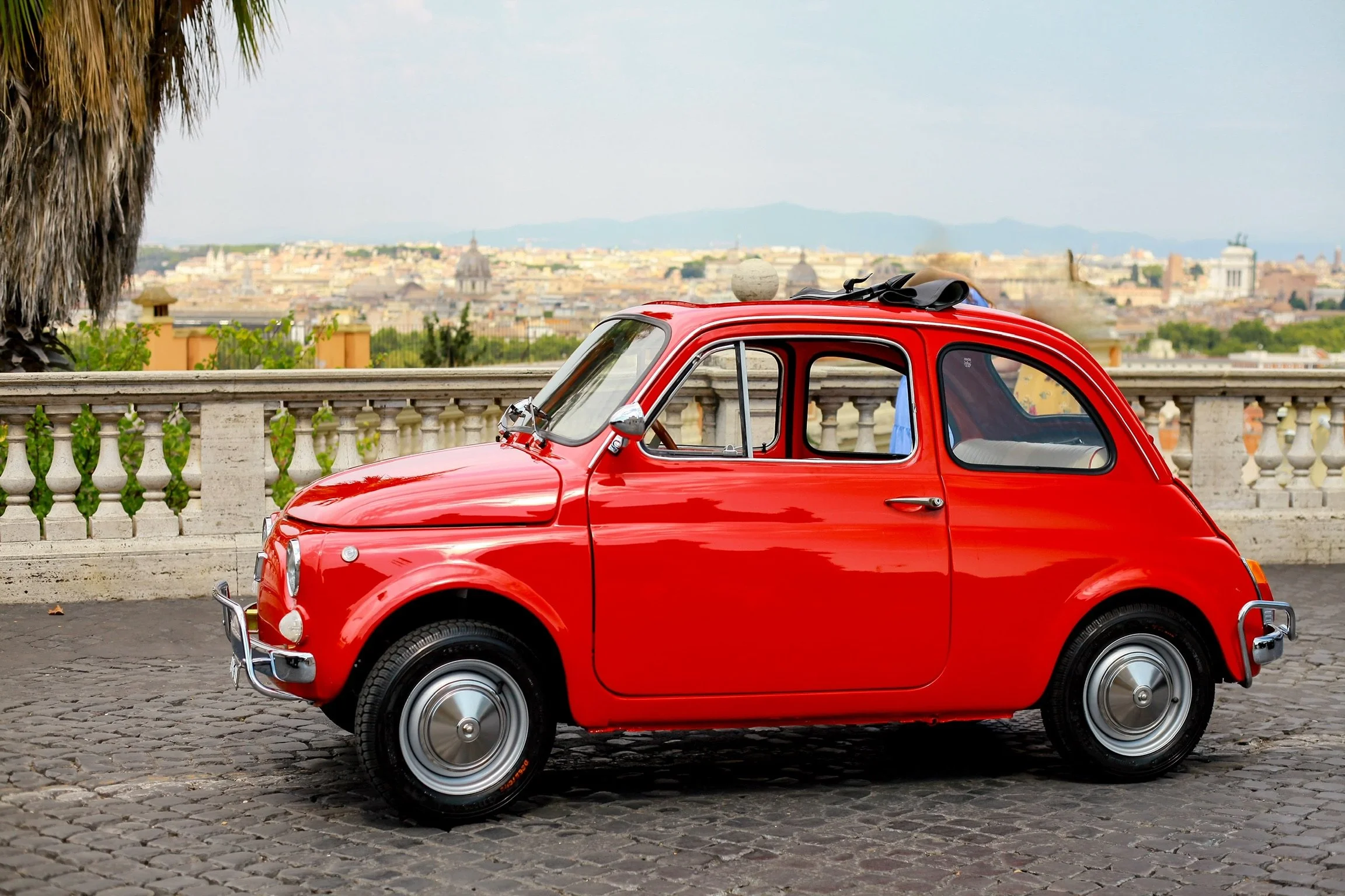 Una vecchia auto rossa Fiat 500 parcheggiata su una strada di ciottoli con un muro di pietra e una città sullo sfondo in un paesaggio urbano con colline e edifici storici.