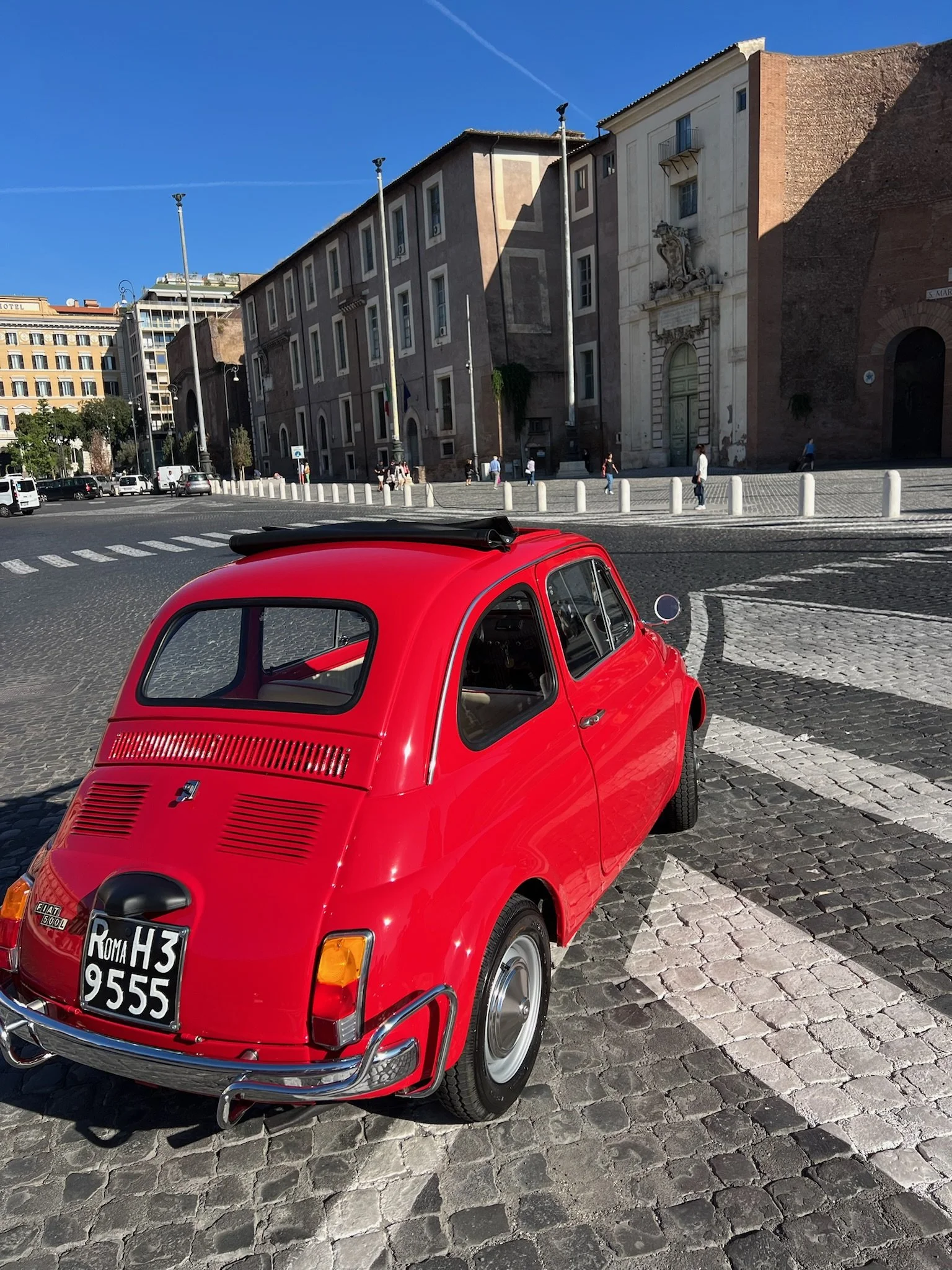 Auto rossa in stile rétro parcheggiata su una strada di ciottoli con edifici storici sullo sfondo, cielo azzurro.