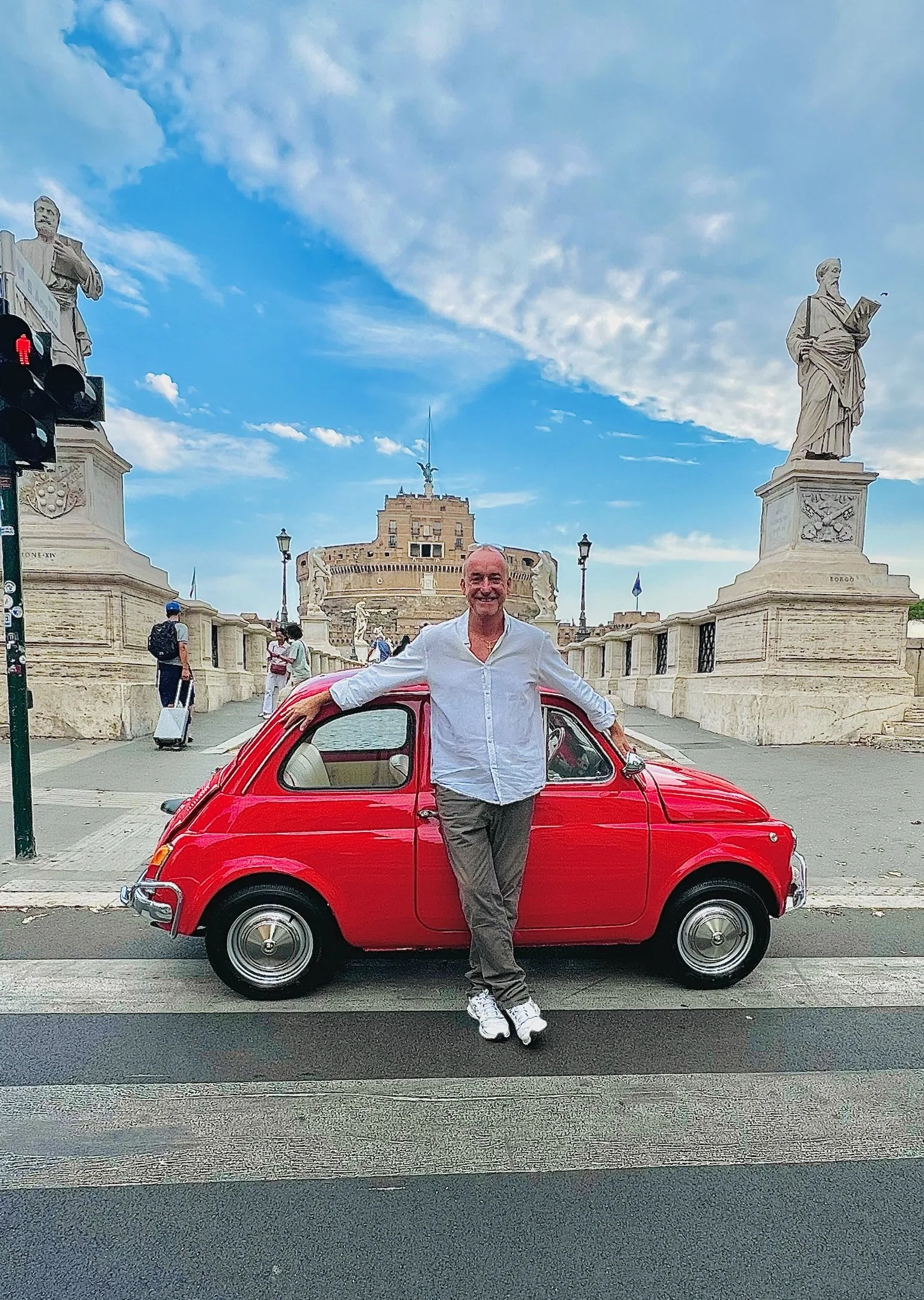 Un uomo sorridente posa con le braccia aperte accanto a una piccola auto rossa sul ponte Sant'Angelo con il Castel Sant'Angelo sullo sfondo a Roma, cielo sereno con alcune nuvole.