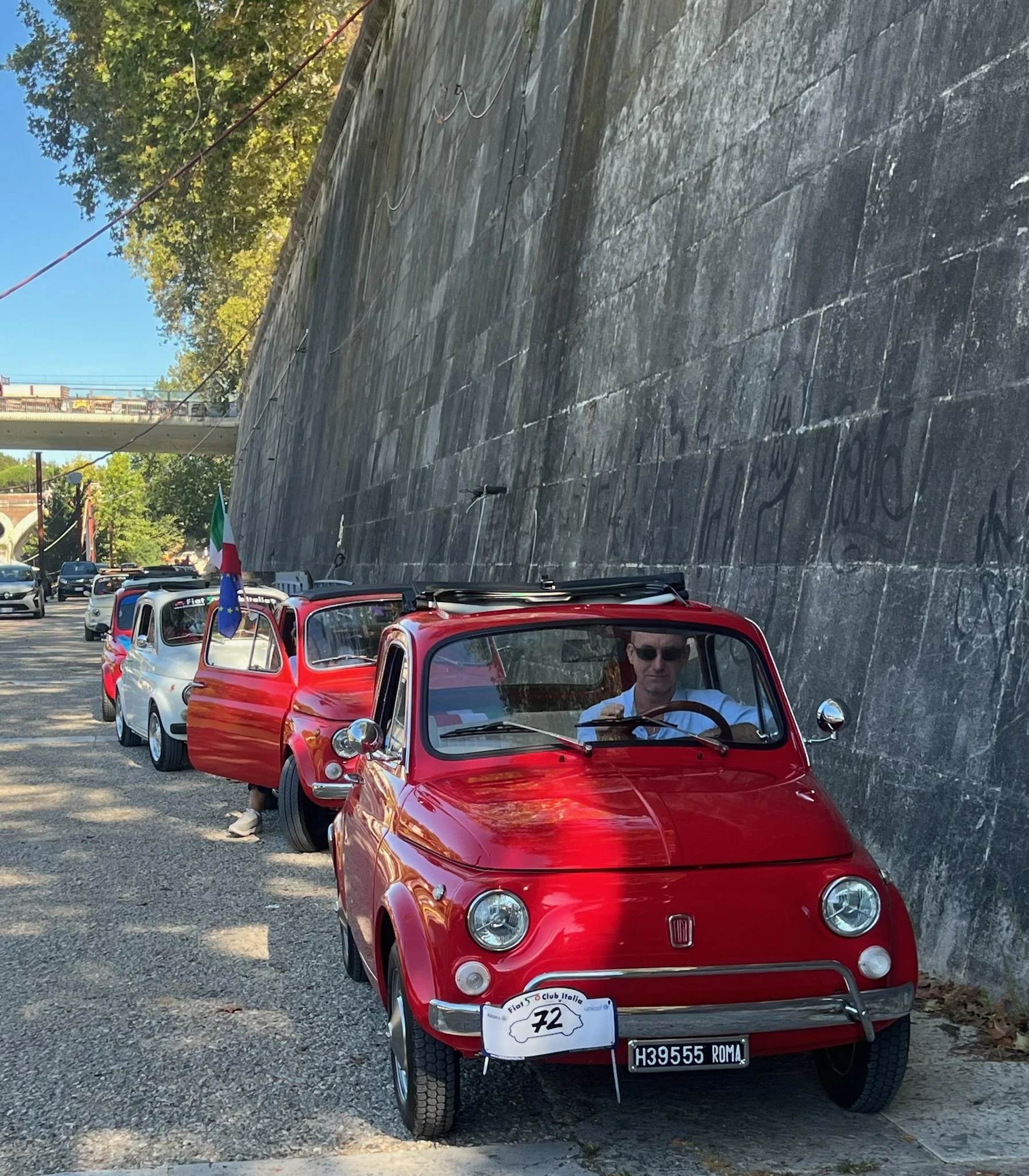 Una fila di piccole auto d'epoca, con la prima vettura rossa in primo piano e un uomo con occhiali al volante, parcheggiate lungo un muro di pietra.