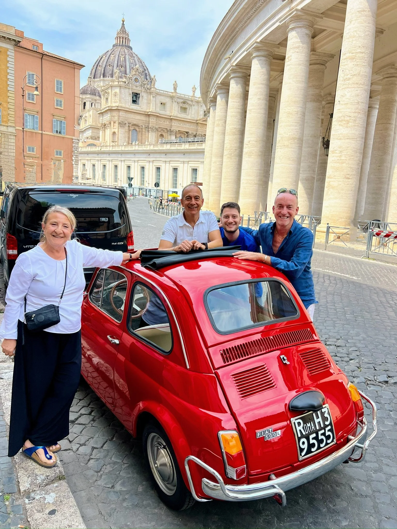 Gruppo di persone che sorridono vicino a una piccola auto rossa Fiat 500 in un'area urbana con il Colosseo in background.