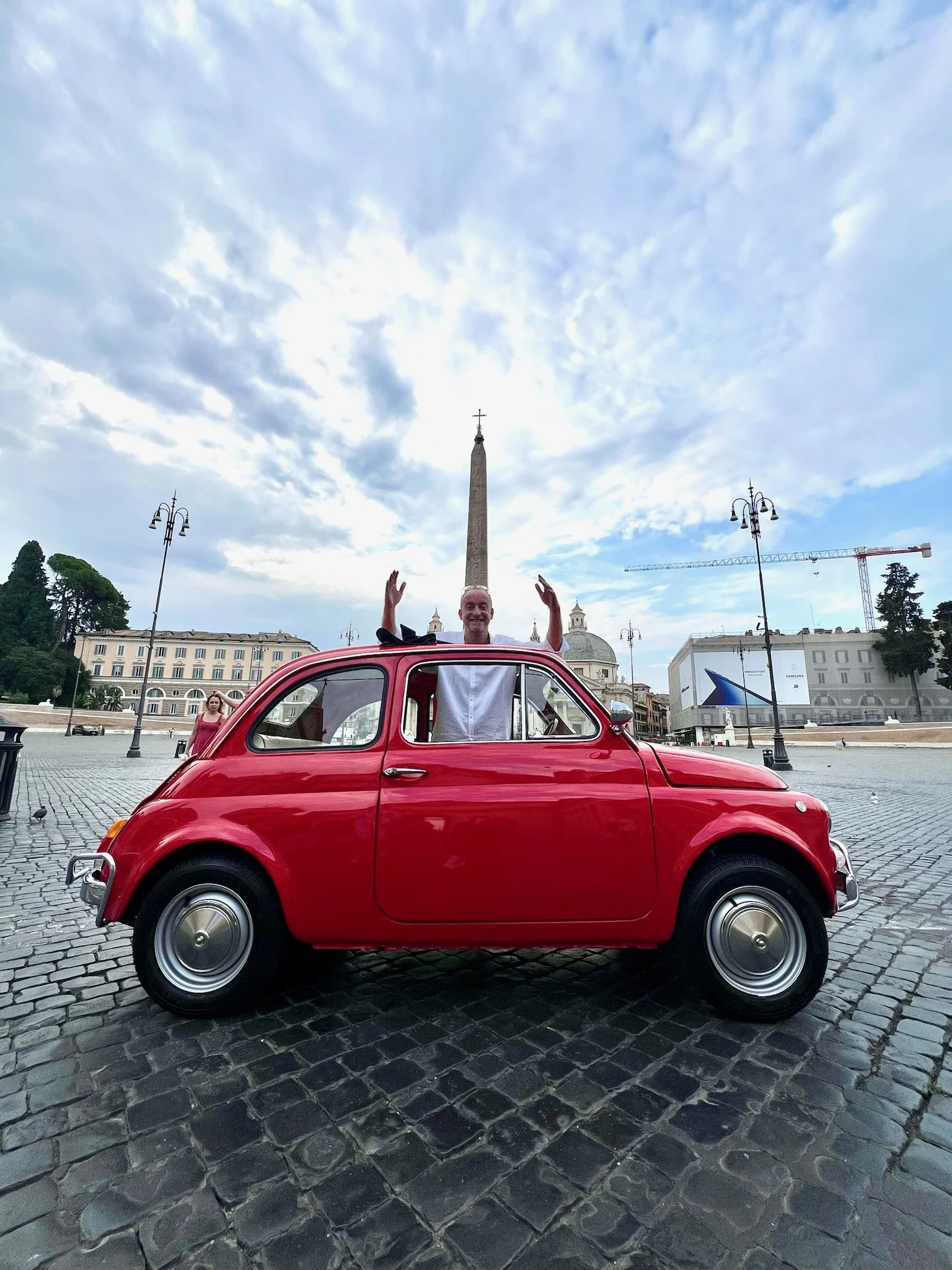 Uomo sorridente che saluta dalla finestra di una macchina rossa vintage, con una piazza e un obelisco sullo sfondo.