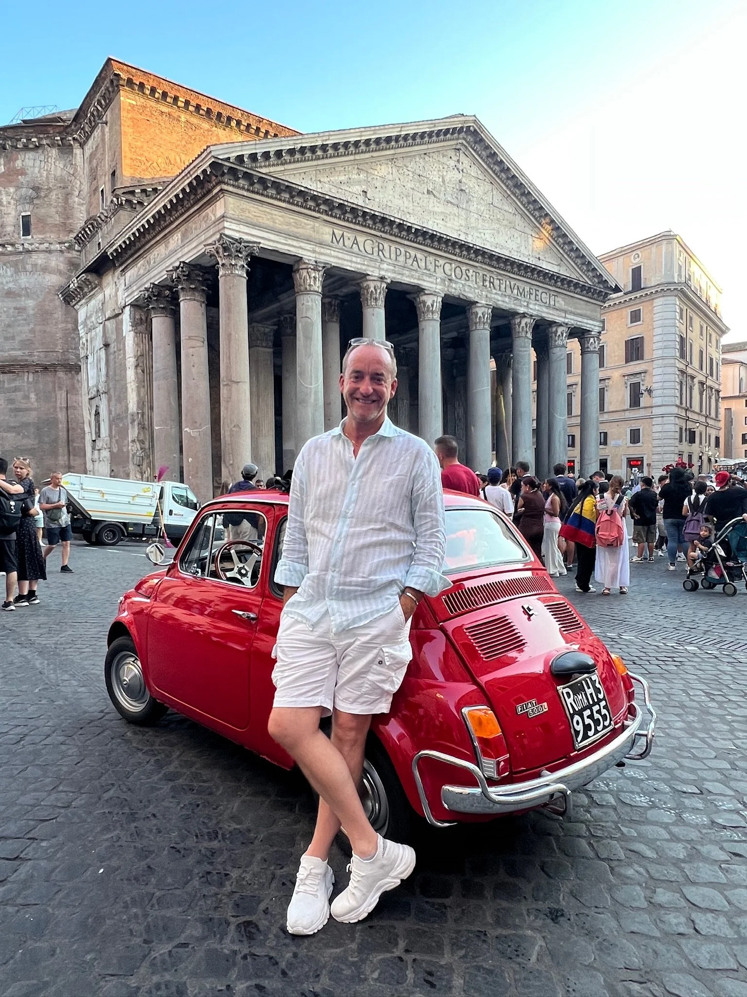 Uomo sorridente in abbigliamento bianco, con scarpe bianche, che si appoggia a una piccola auto rossa Fiat 500, con un monumento storico e molte persone sullo sfondo a Roma.