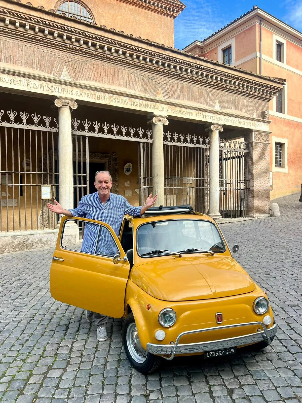 Uomo sorridente con occhiali sulla testa, indossa una camicia blu a quadri, si appoggia su una piccola auto gialla con porta aperta, di fronte a un edificio storico con colonne e dettagli architettonici antichi, sullo sfondo un cielo blu con alcune n